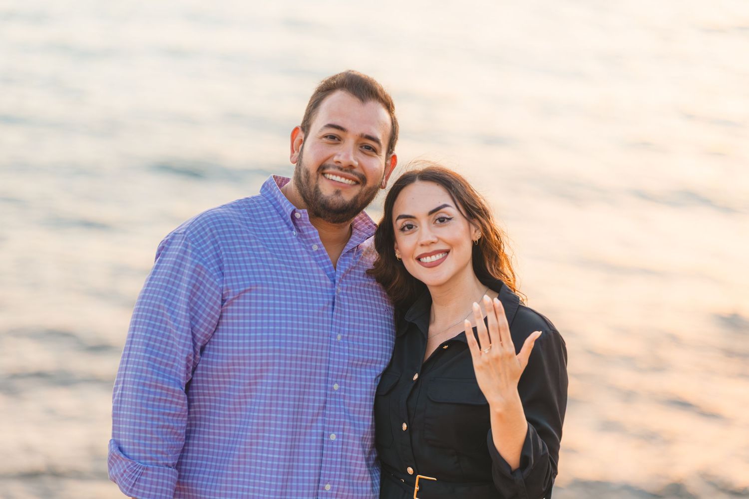 A couple smiles together on a coastal cliff during sunset with the Pacific Ocean in the background.