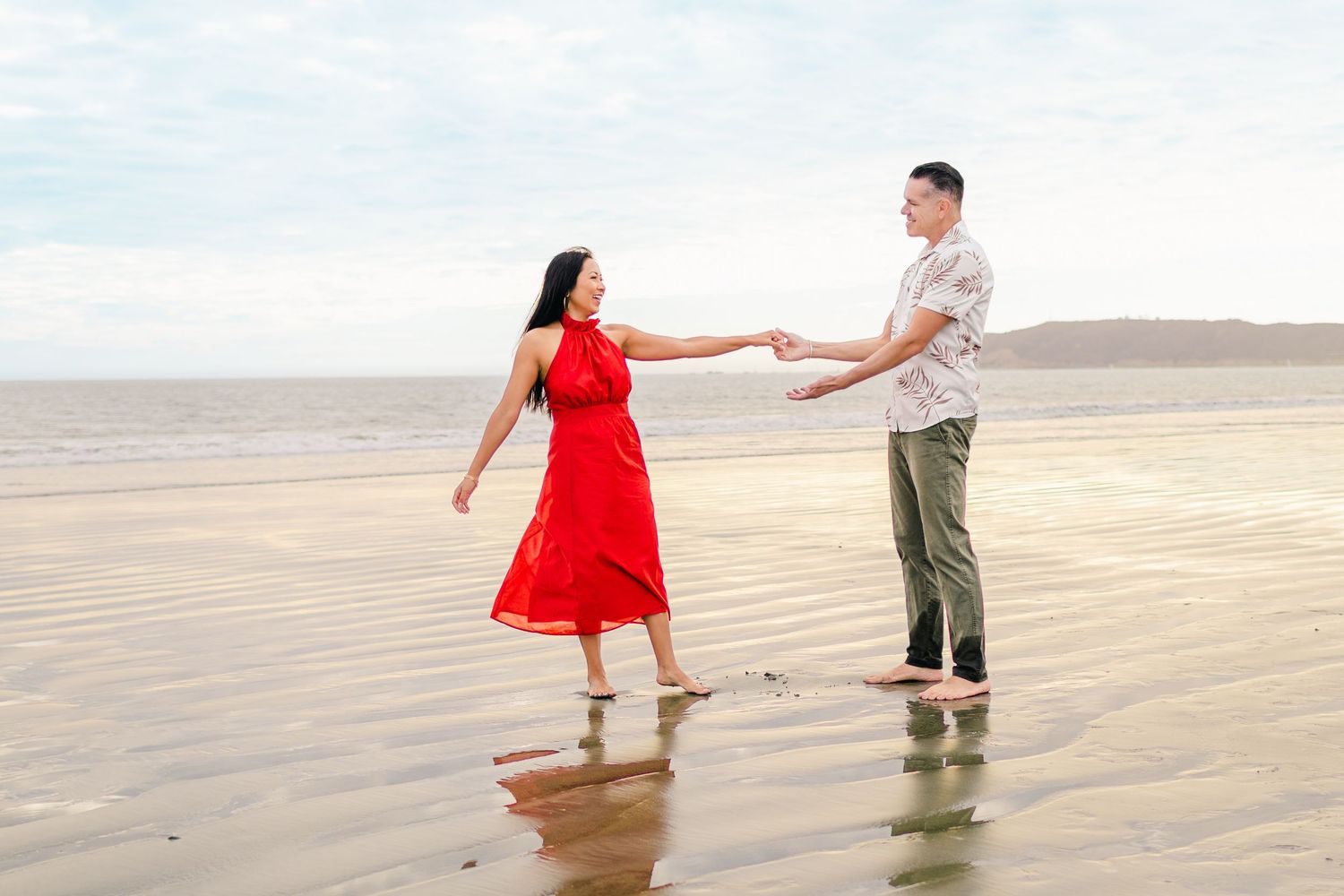 A couple in red dress and casual wear holds hands while walking along a misty beach shoreline.