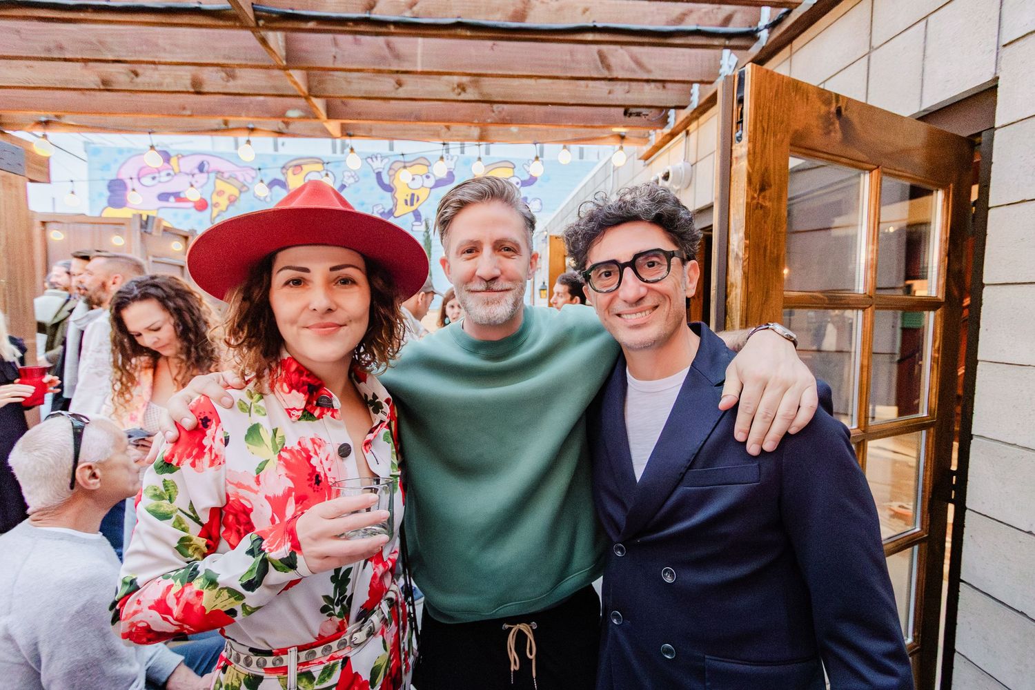 Three people smiling together under wooden pergola at outdoor gathering.