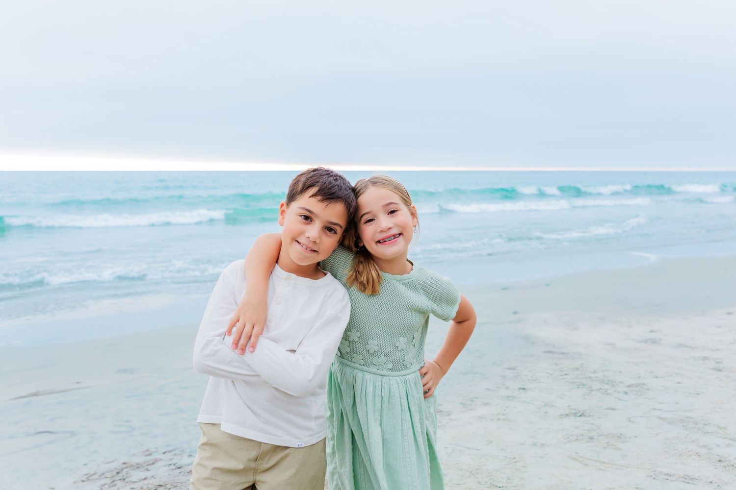 Two children pose together on a beach with turquoise waves rolling in the background.