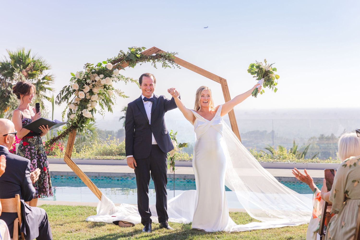 Wedding ceremony setup with wooden arch decorated in white fabric and greenery by the ocean.