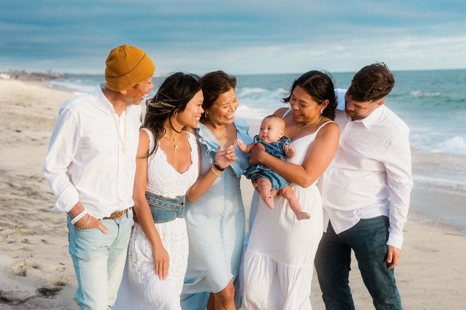 A group gathers around a baby on the beach, all dressed in white and blue coordinated outfits.
