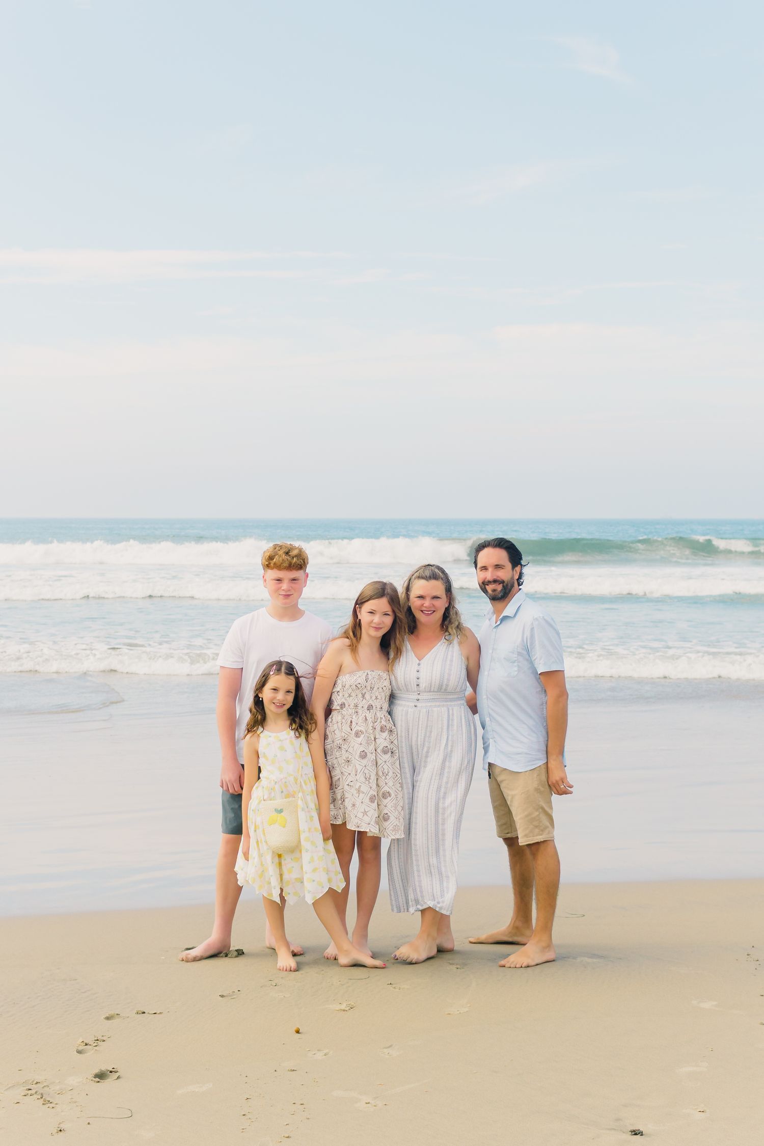 A group in light summer attire poses together on a sandy beach with ocean waves in the background.