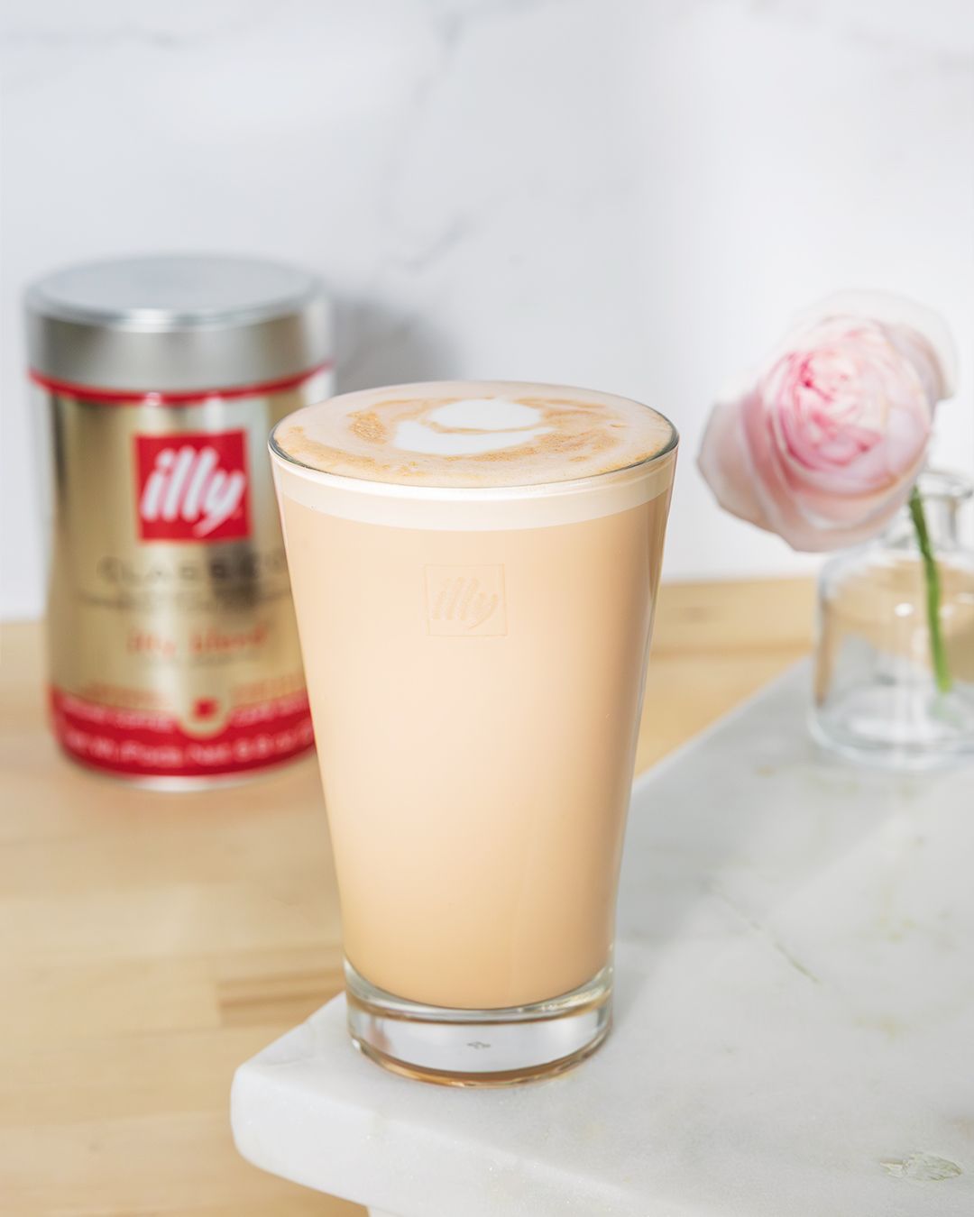 Creamy latte in clear glass with illy coffee tin and pink rose on marble surface.