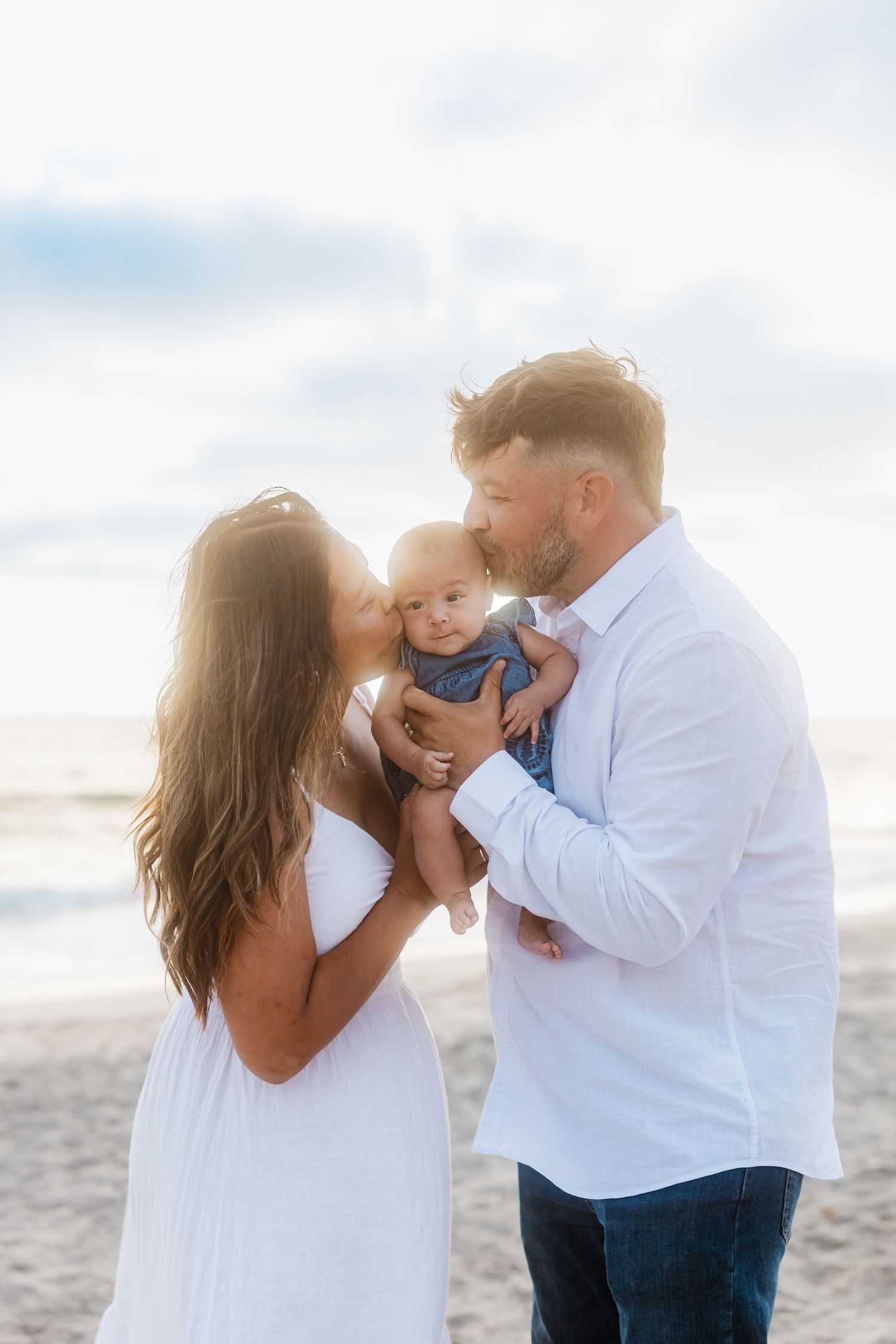 A loving couple shares a tender moment with their baby on a sun-drenched beach at sunset.