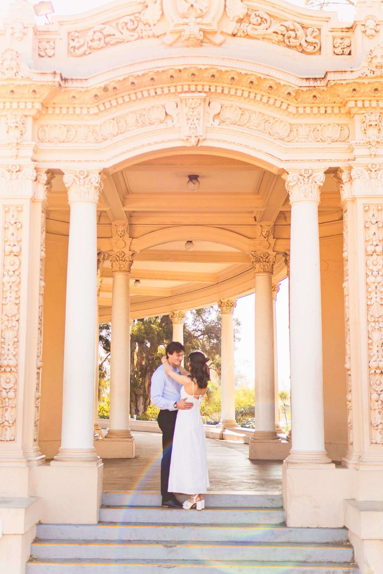 A couple embraces under ornate cream-colored columns and arches in a romantic outdoor setting.