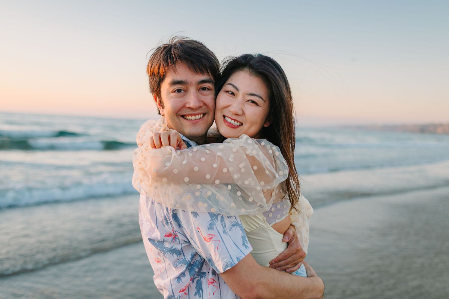 A couple embraces joyfully on a sunny beach at sunset with ocean waves in the background.
