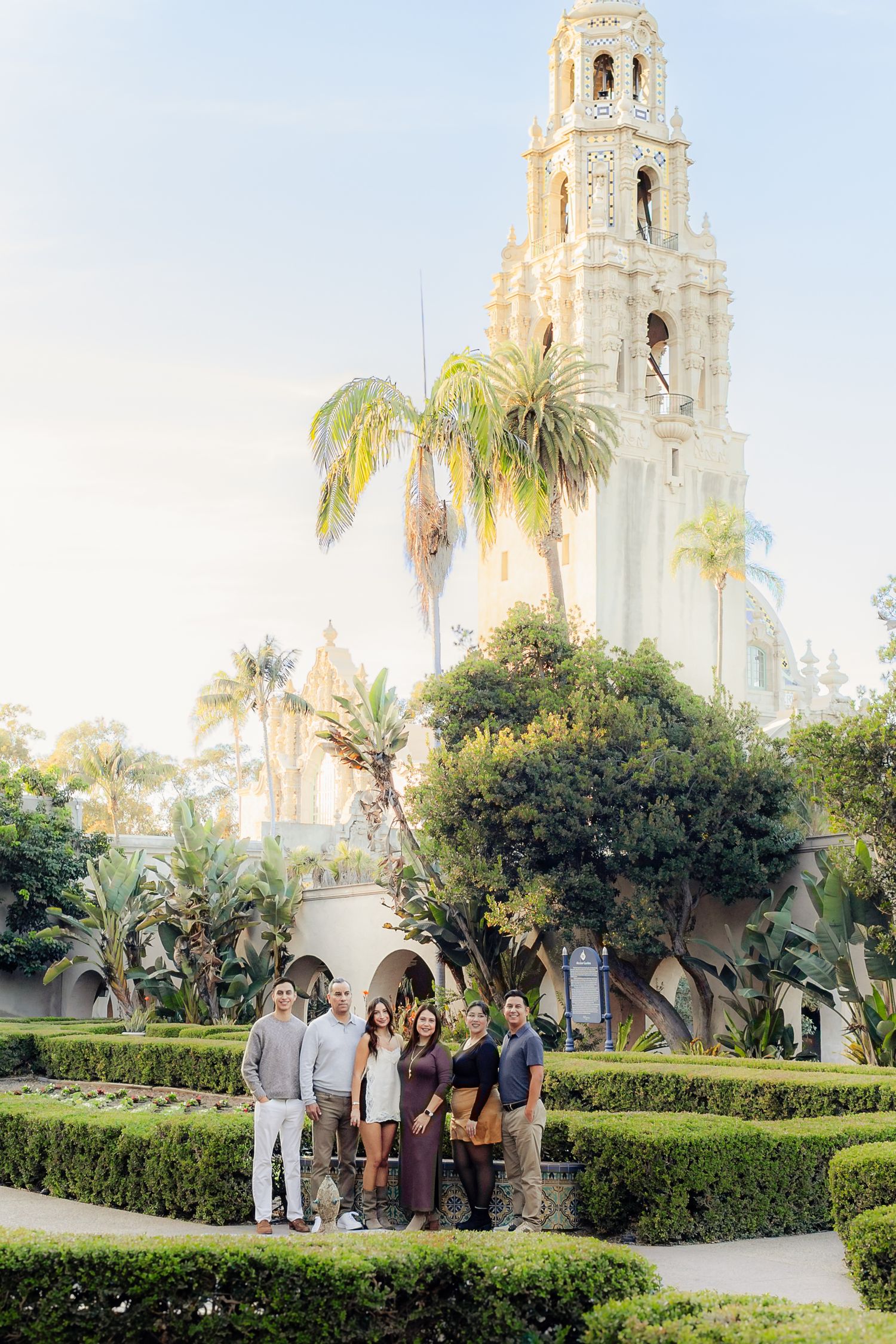 Group of people walking along manicured garden paths at a Spanish colonial style church with white towers and palm trees.
