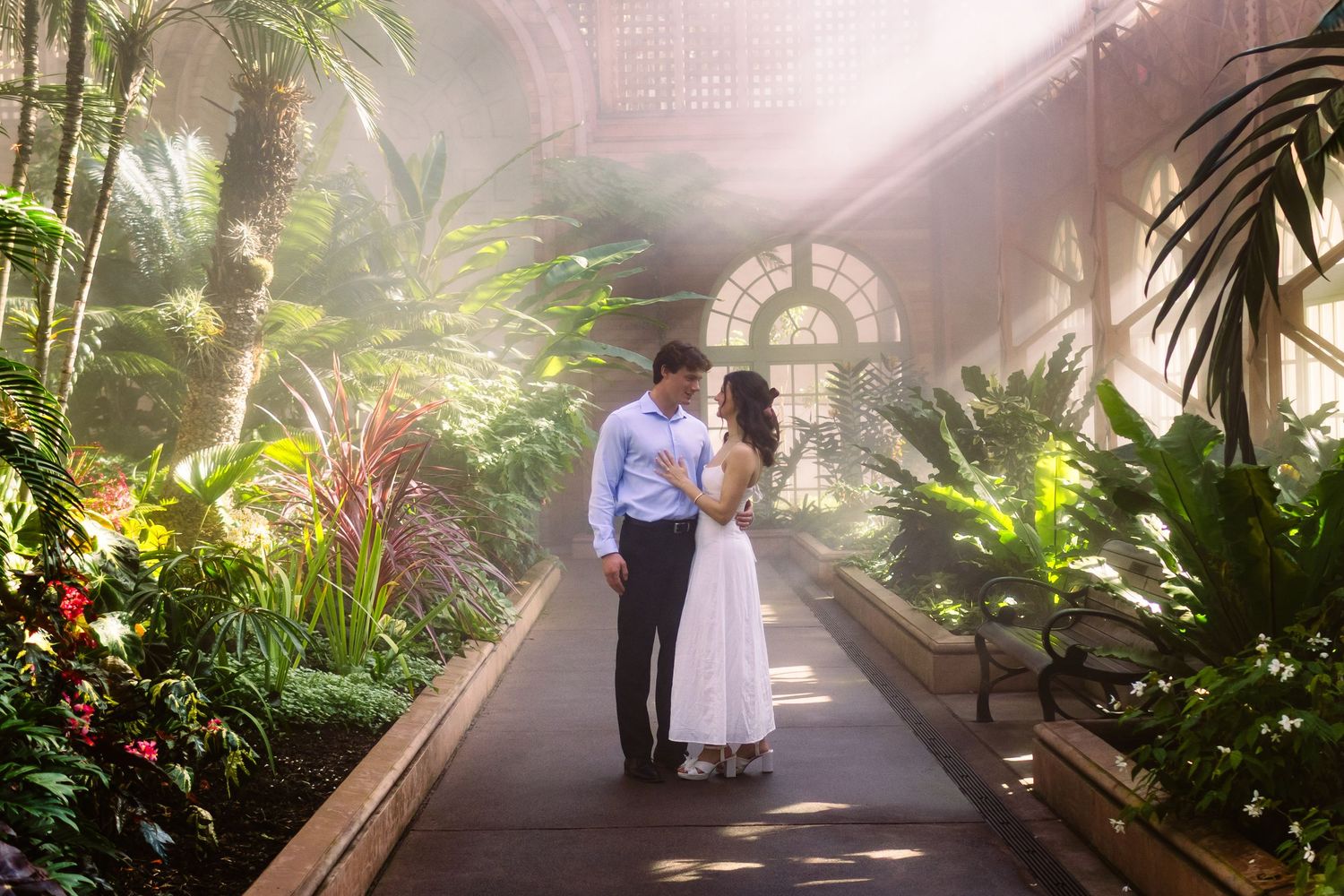 A couple walks through a lush indoor garden conservatory filled with tropical plants and misty sunlight.