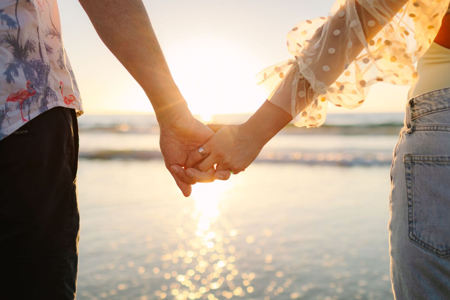 A couple holds hands on a beach at sunset with golden sunlight sparkling on the ocean waves in the background.
