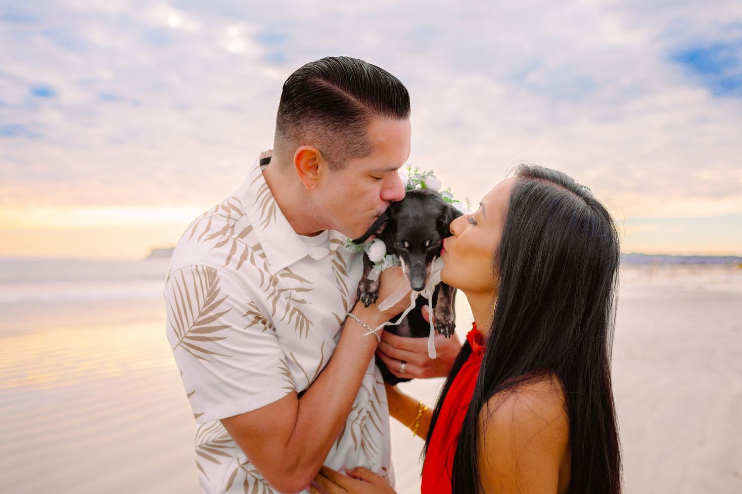 A couple in tropical attire shares romantic moments on a beach at sunset with the ocean in the background.
