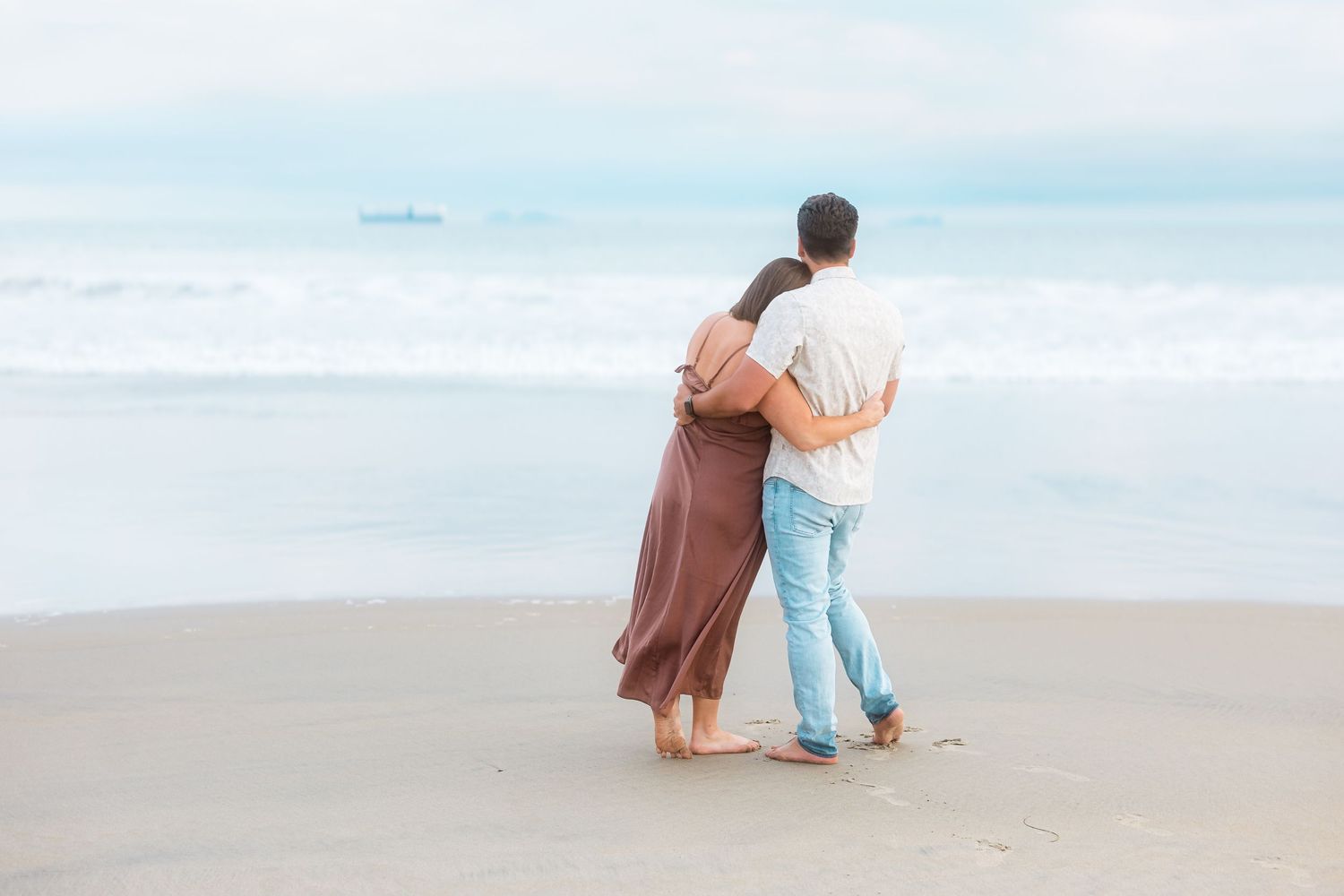 Couple walks arm in arm along misty beach shore during maternity photoshoot.