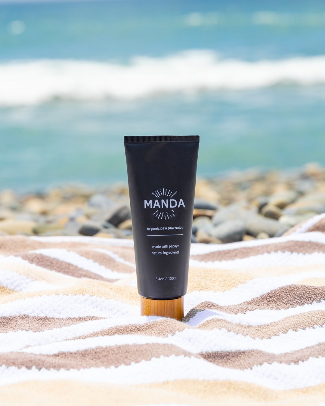 Black Manda skincare tube against blurred ocean background on sandy beach.
