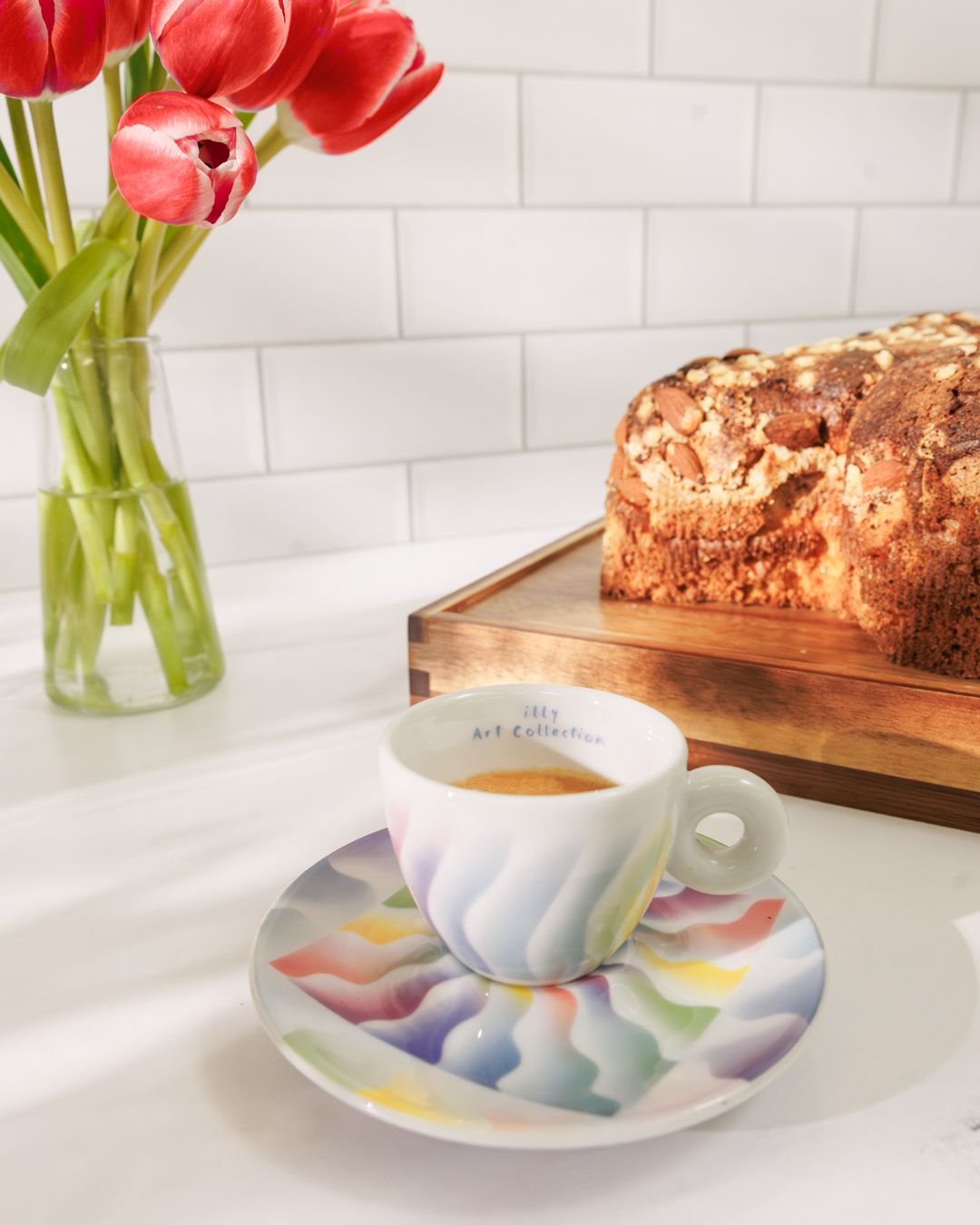 Coffee cup on colorful saucer with red tulips and fresh bread in background.