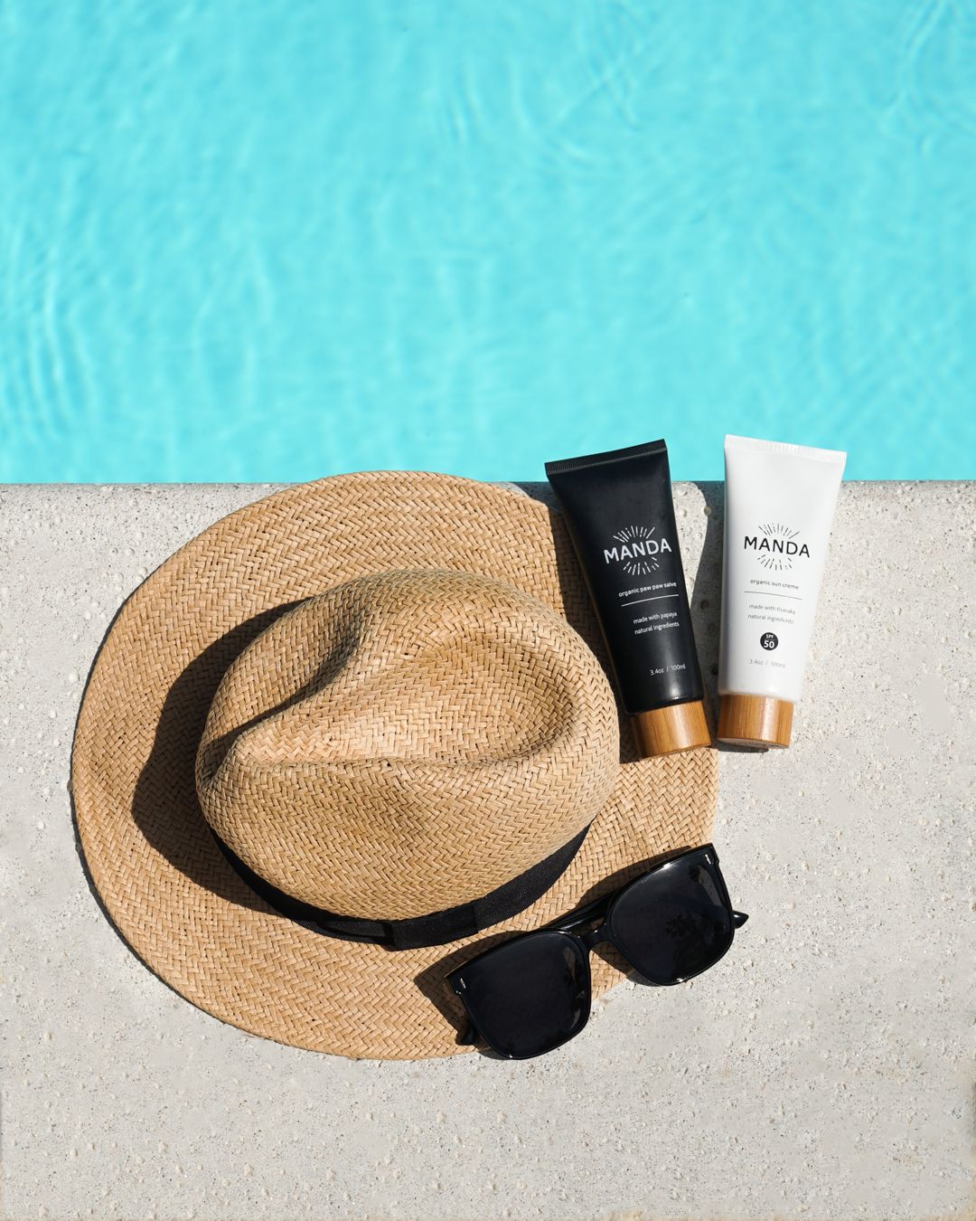 Straw hat and sunglasses with Manda skincare products by poolside.