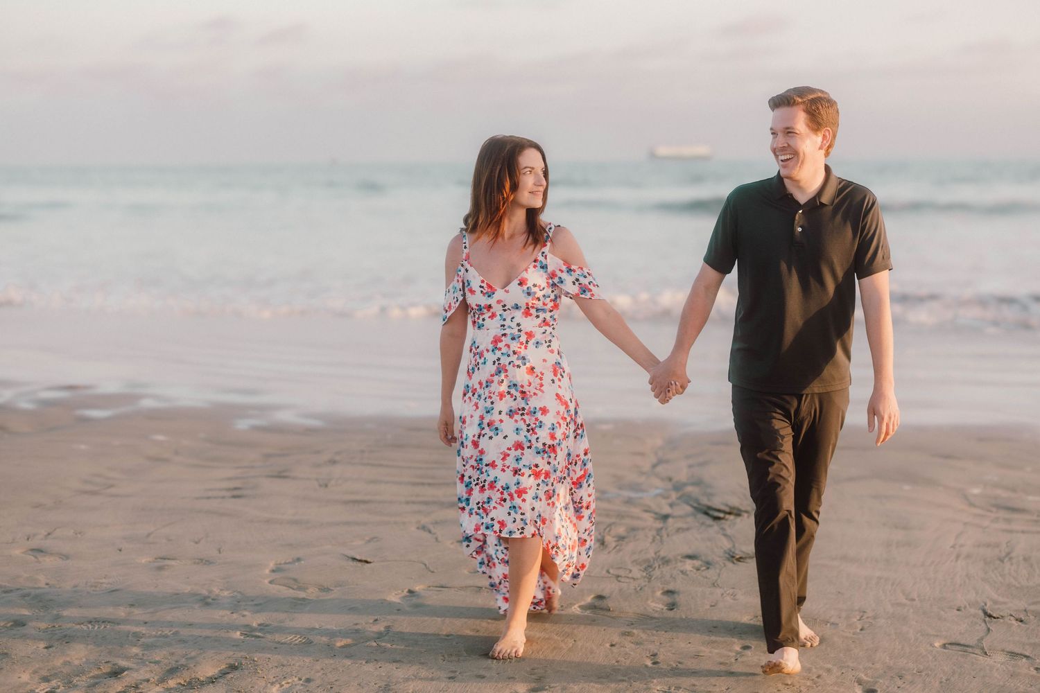Couple walking and laughing together along peaceful beach shore during sunset photo session.