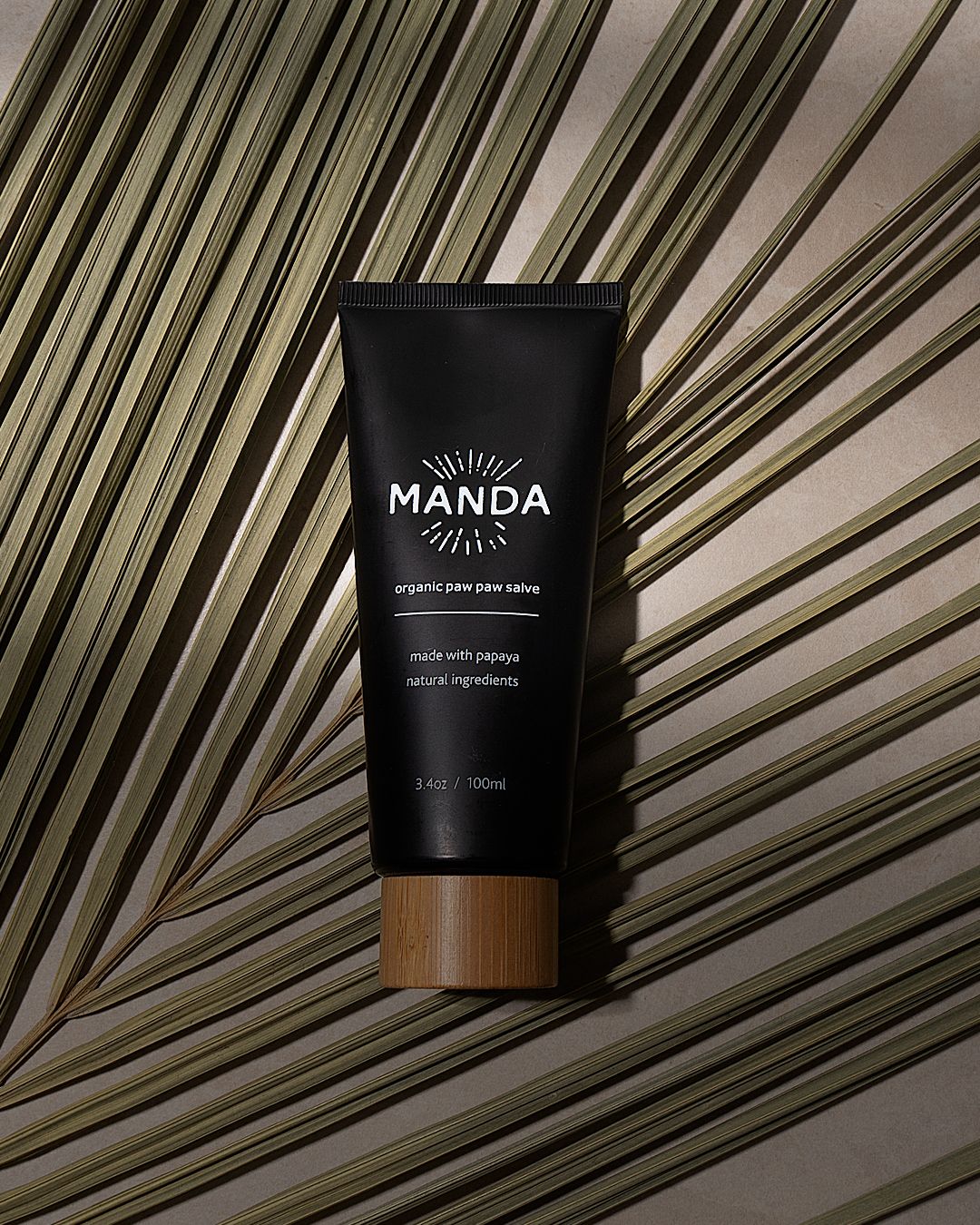 Manda black skincare tube with bamboo cap photographed against striped palm leaf shadows.