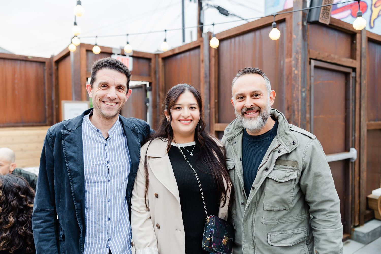 Three people stand smiling together outside a rustic restaurant with wood paneling and string lights.