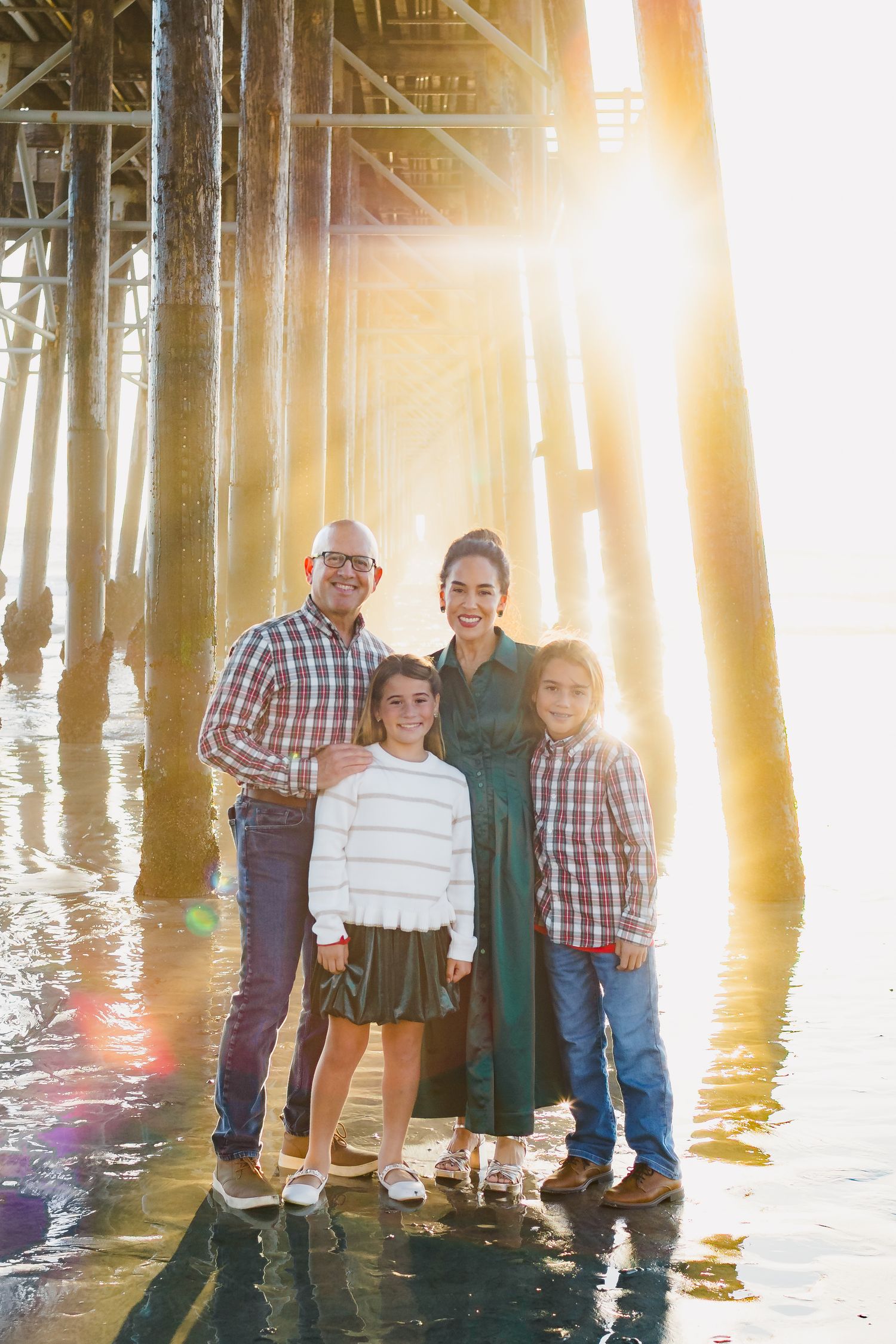 Family portrait under wooden pier with sunlight streaming through posts creating golden glow at sunset.