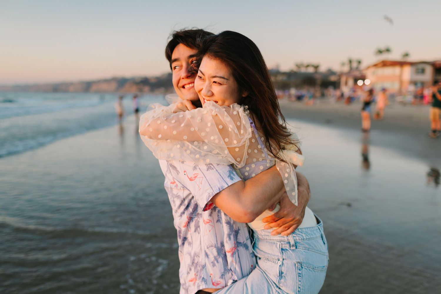 A joyful embrace between two people on a beach at sunset with ocean waves and beachgoers in the background.