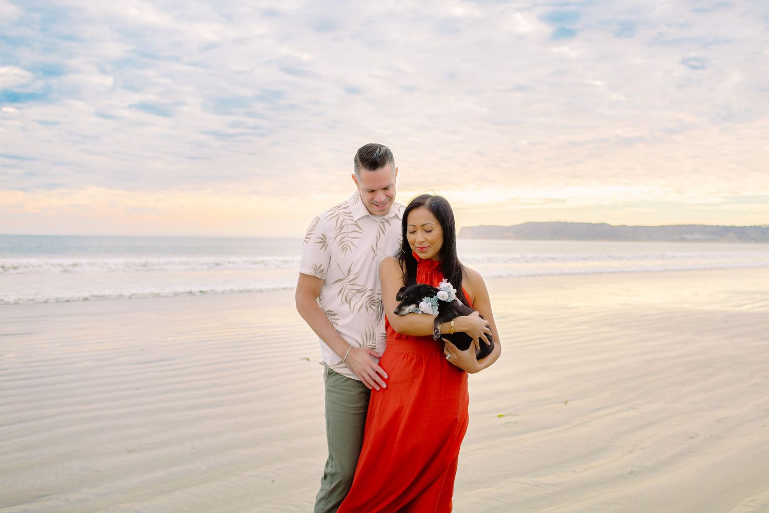 A couple in red and white outfits walks together on a sandy beach during sunset with waves in the background.