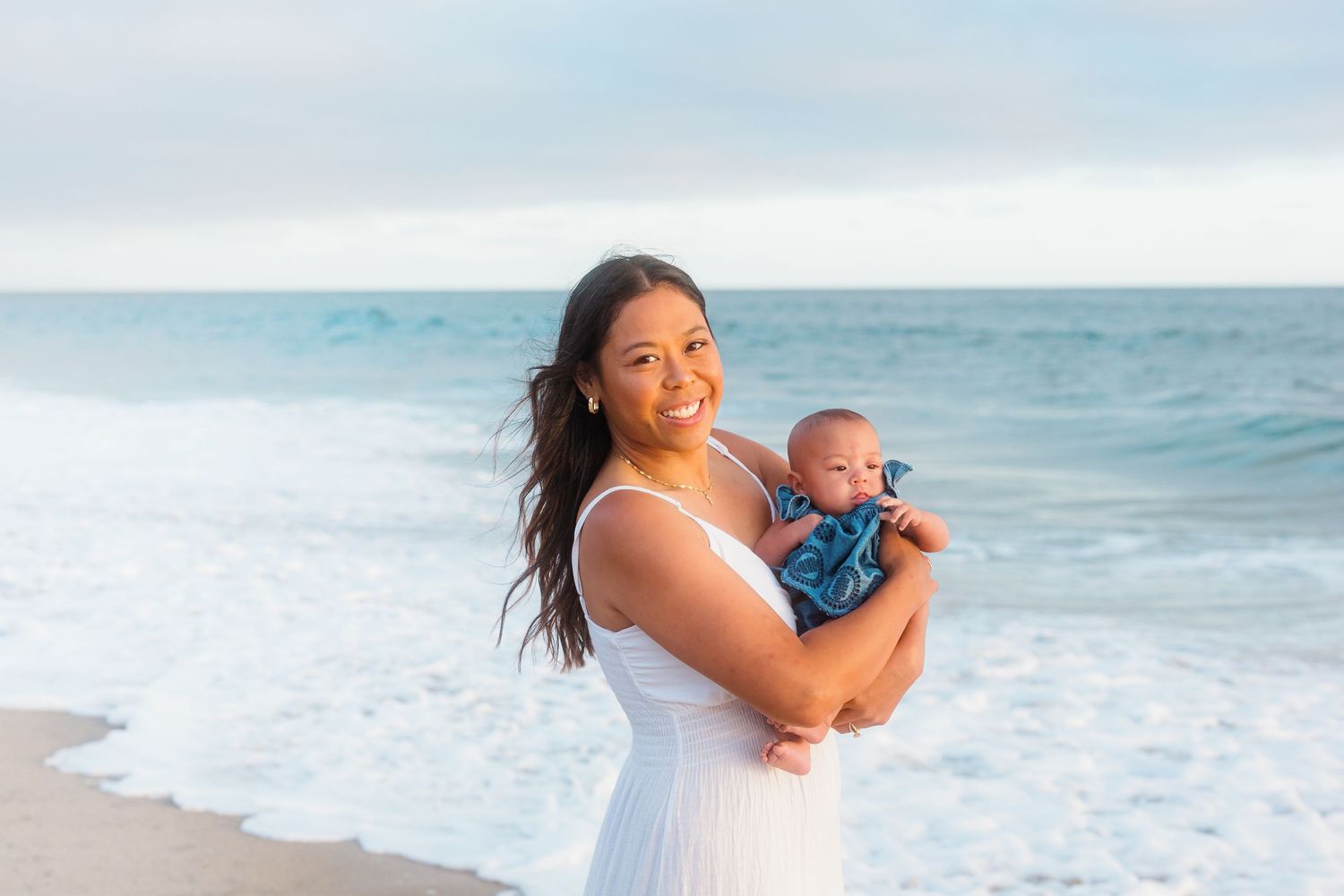A woman in white holding a baby poses against the ocean backdrop.