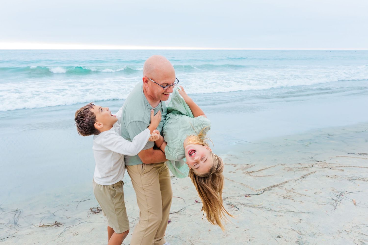 Playful family moment captured on the beach with ocean waves behind them.