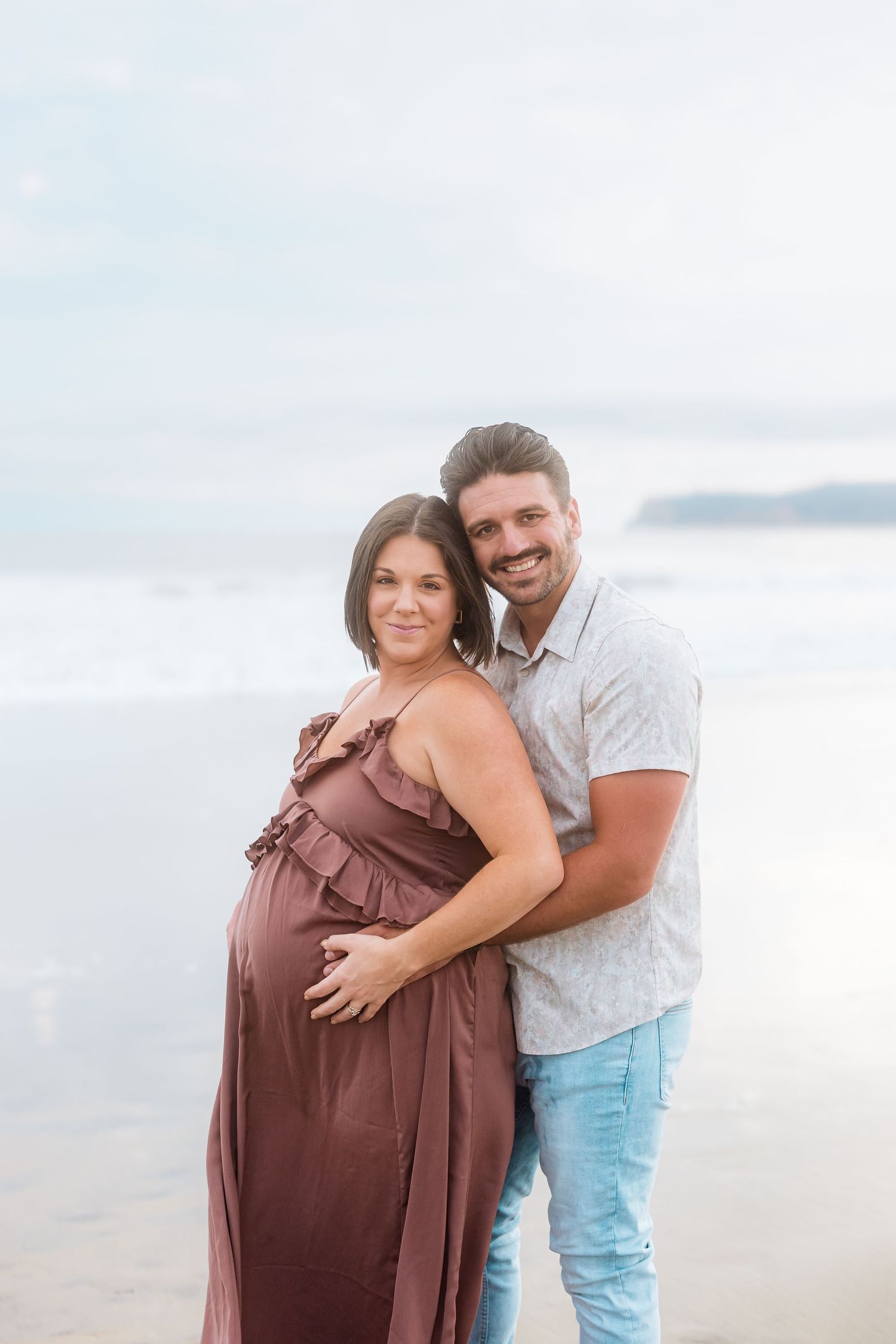 Couple embraces on beach at sunset with pregnant woman wearing flowing mauve dress and man in white shirt.