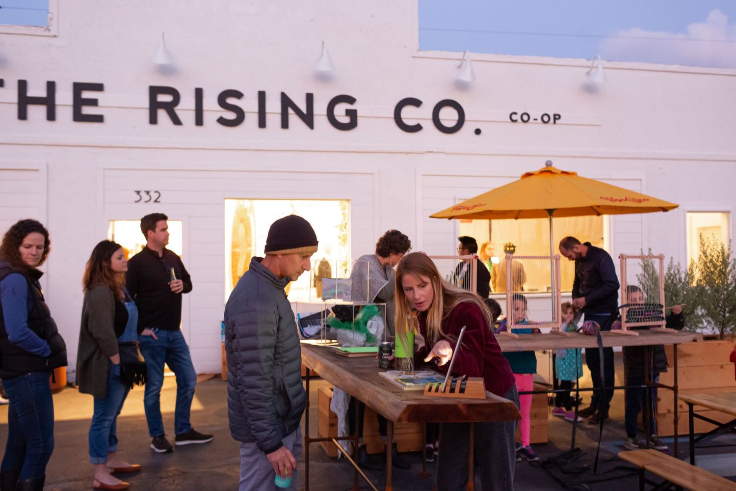 People gather outside The Rising Co retail store at dusk with string lights illuminating the storefront area.