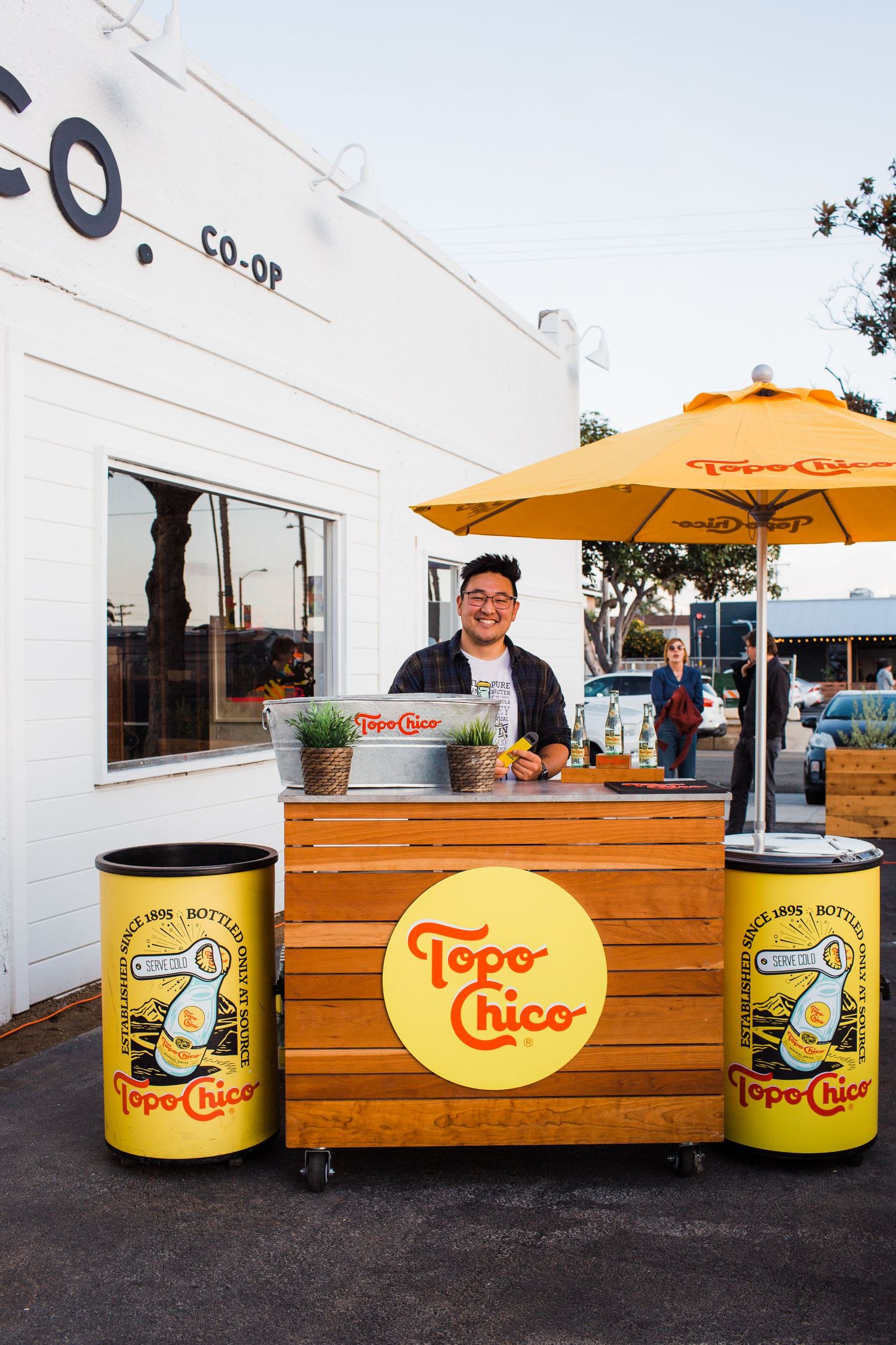 Yellow and orange Topo Chico retail stand with wooden countertop and umbrellas set up outdoors during daytime.