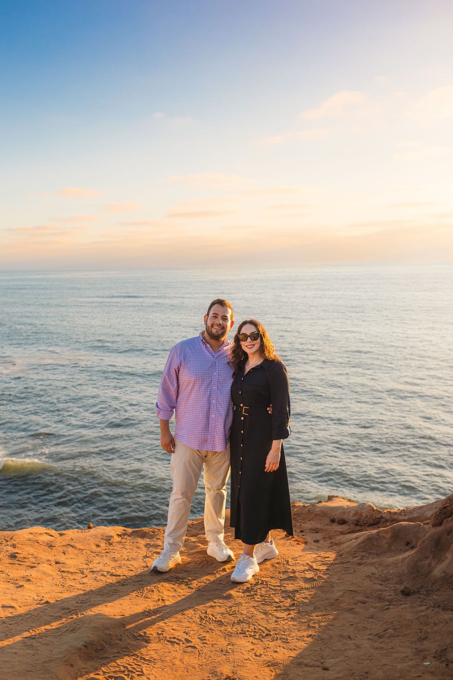 A couple stands on a rocky coastal cliff during golden hour with the ocean and sunset in the background.