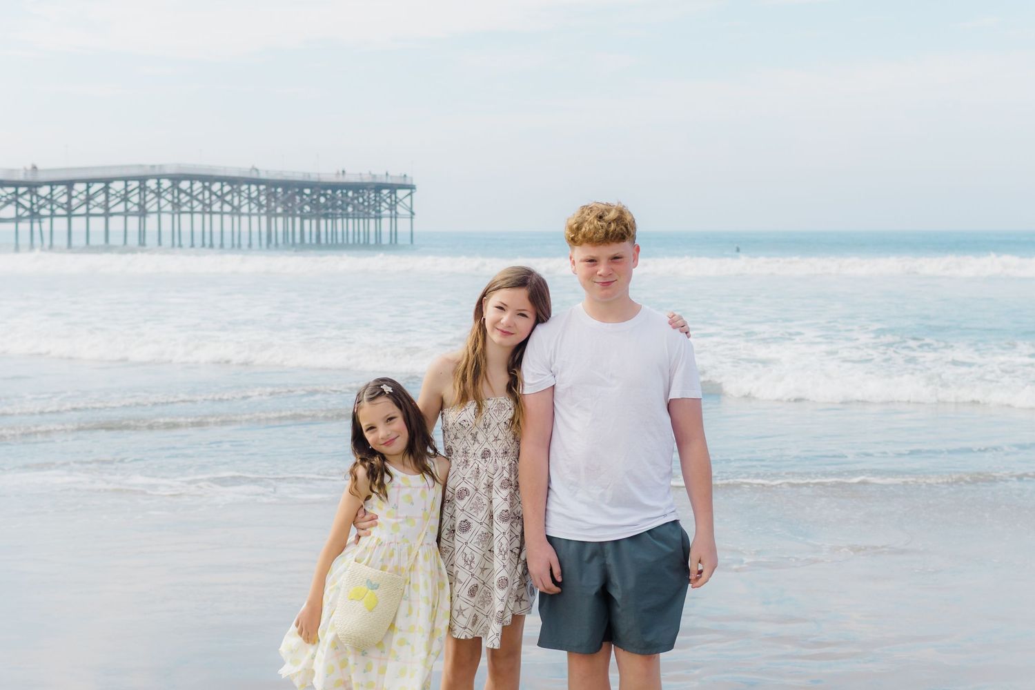 Three people stand together on a beach with a wooden pier in the background on a sunny day.