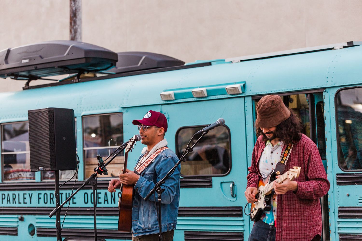 Musicians perform with guitars next to a turquoise vintage bus during an outdoor event.