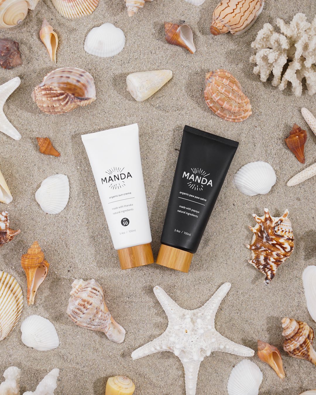 Manda black and white skincare tubes arranged with seashells and starfish on sand.