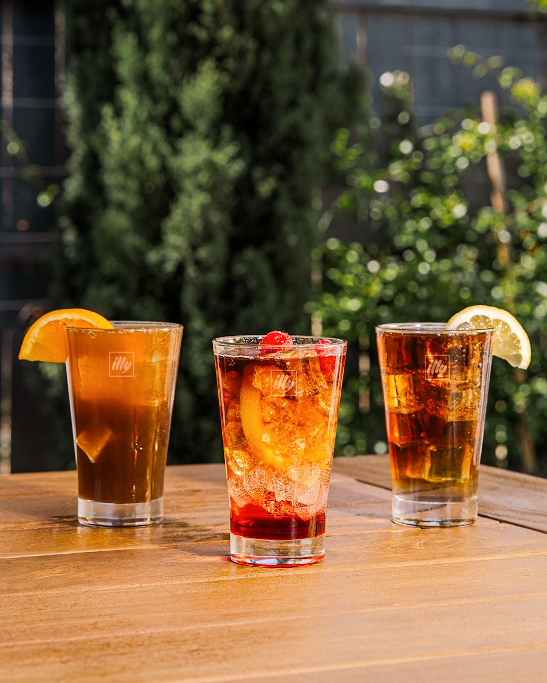 Row of amber-colored cocktails on wooden table with outdoor garden view.