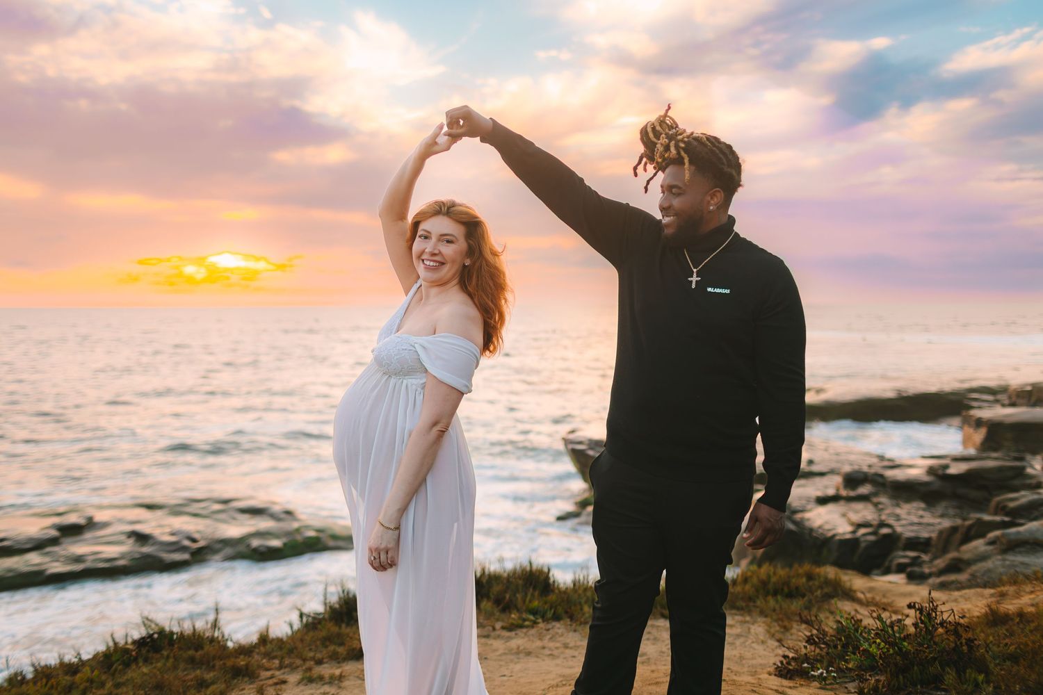 A couple in black and white attire embraces on a rocky beach at sunset with ocean waves crashing behind them.