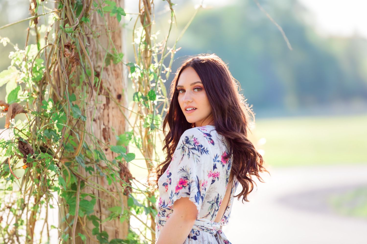 Woman in floral dress poses against vine-covered wall in outdoor garden setting.