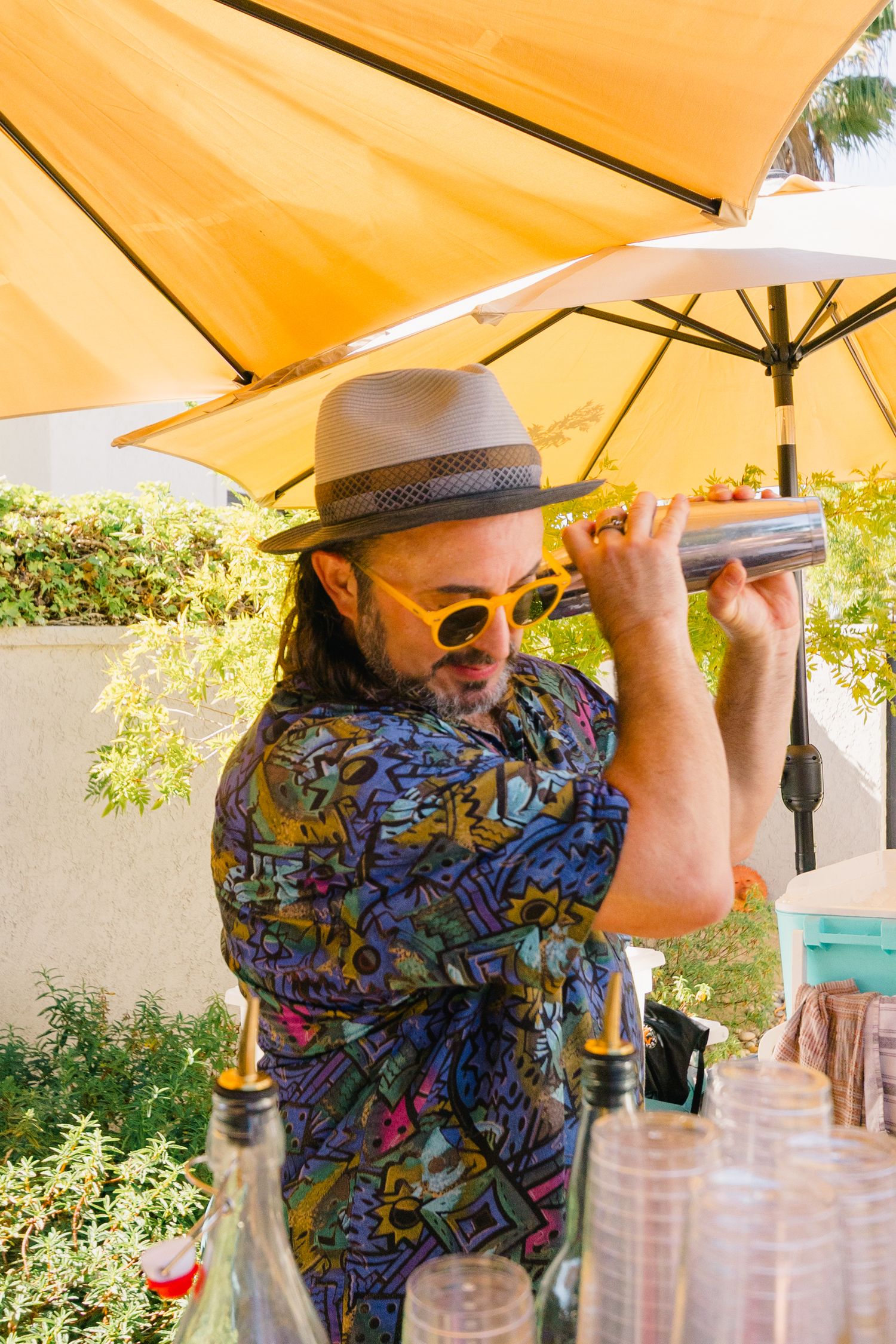 Someone wearing a colorful patterned shirt pouring drinks under a yellow umbrella.