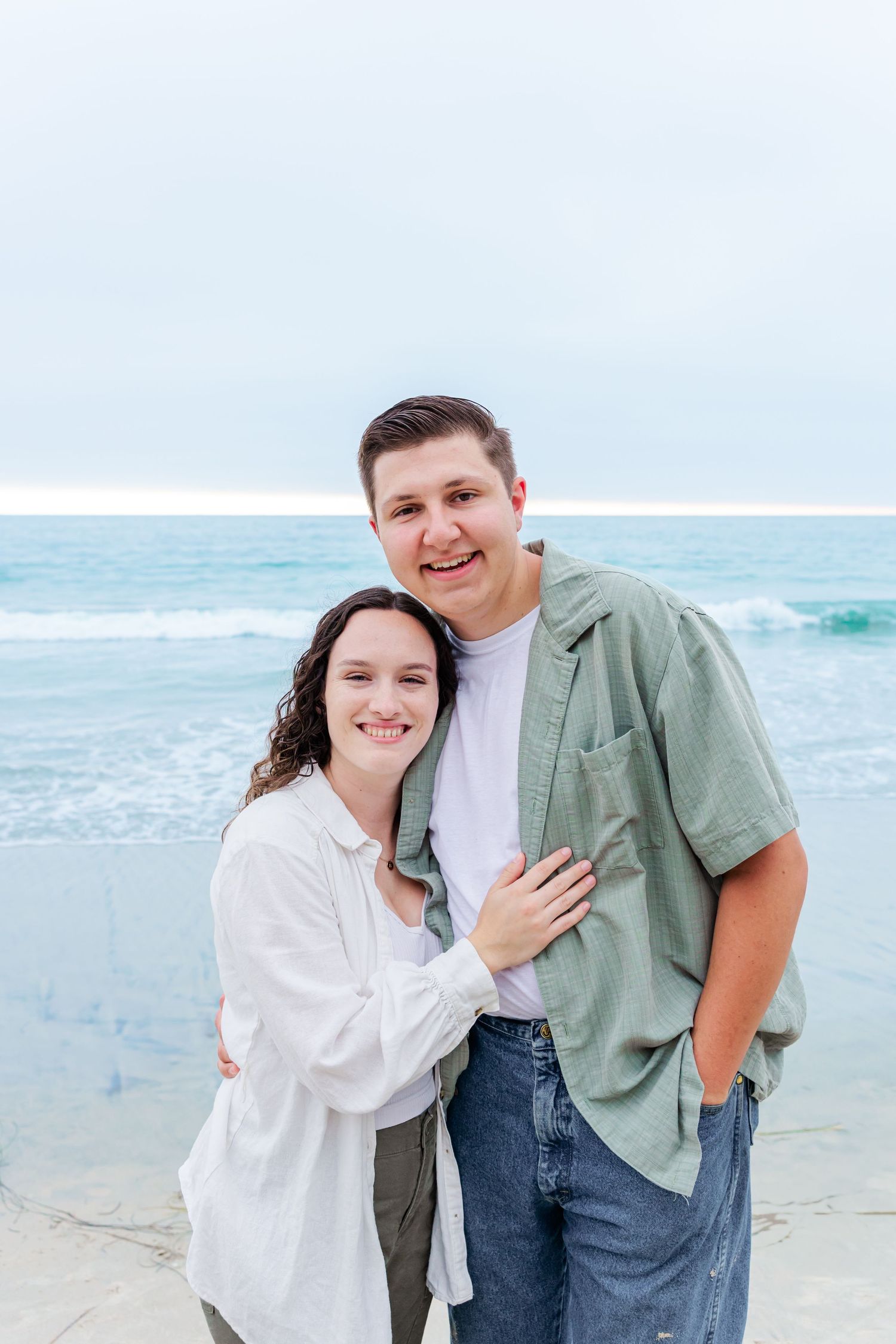 A couple embraces while posing for a casual beach photo with the ocean behind them.