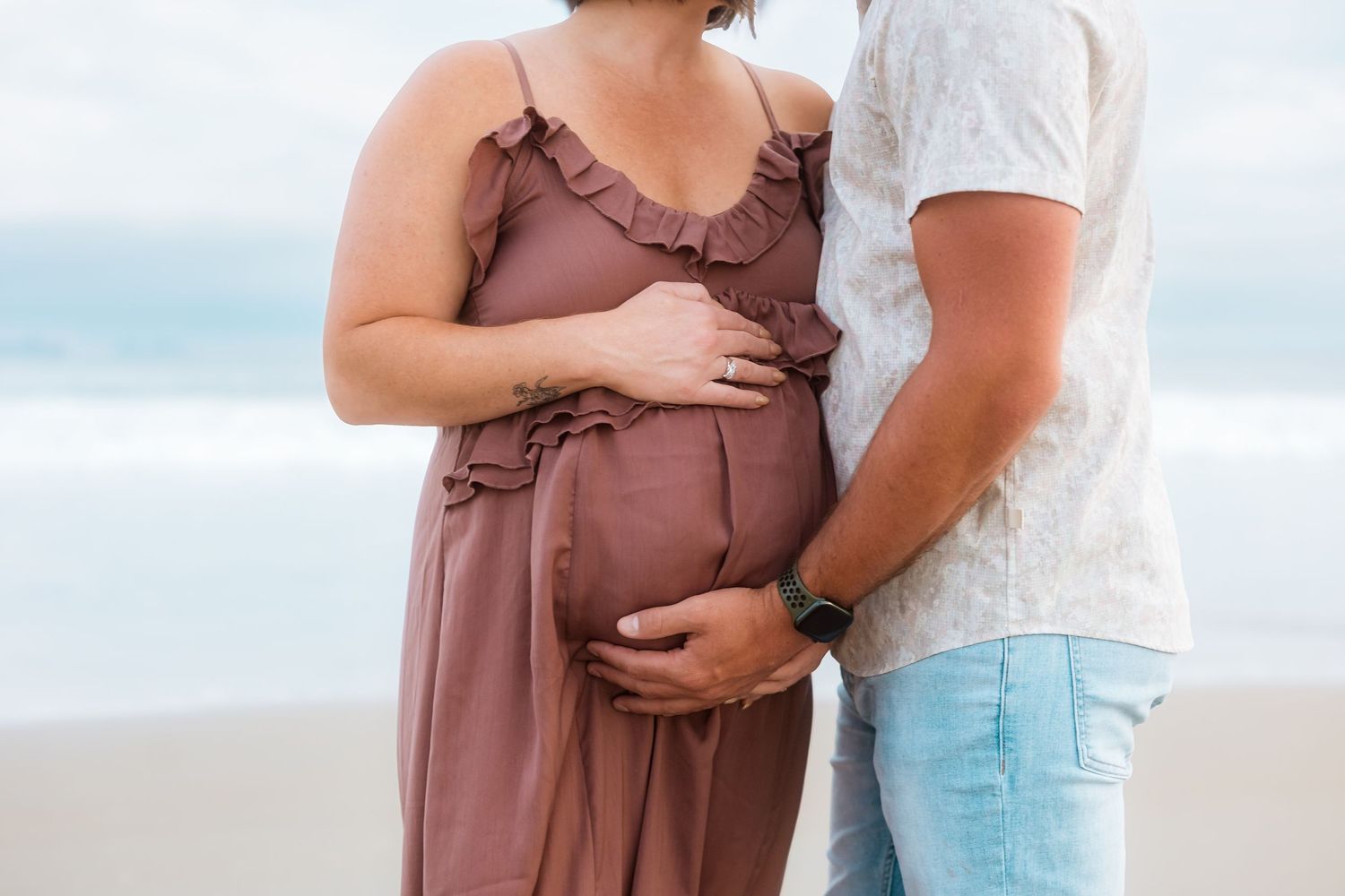 Close up of expectant parents' hands cradling baby bump in ruffled mauve dress.