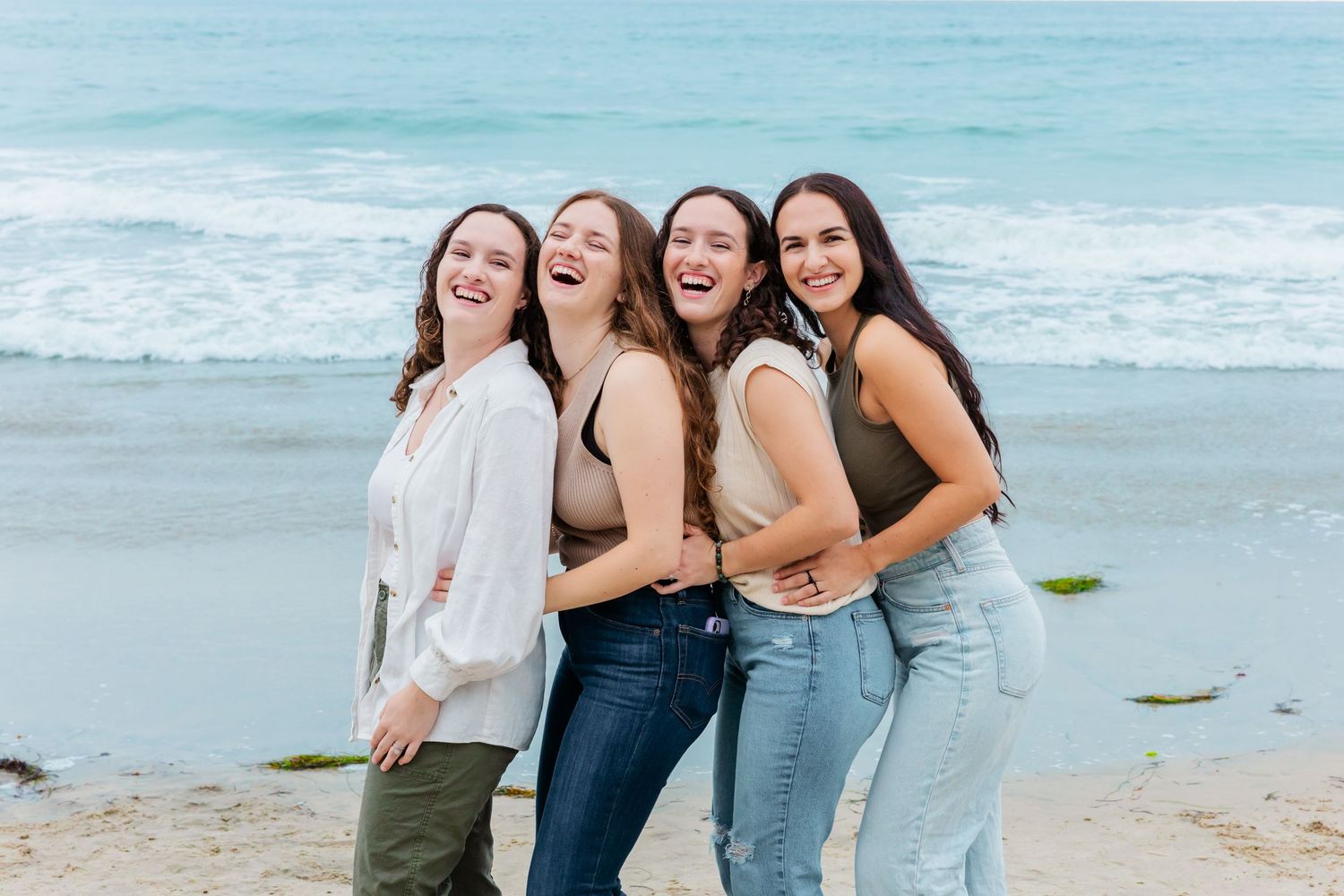Four friends laugh together while posing on a beach with ocean waves behind them.