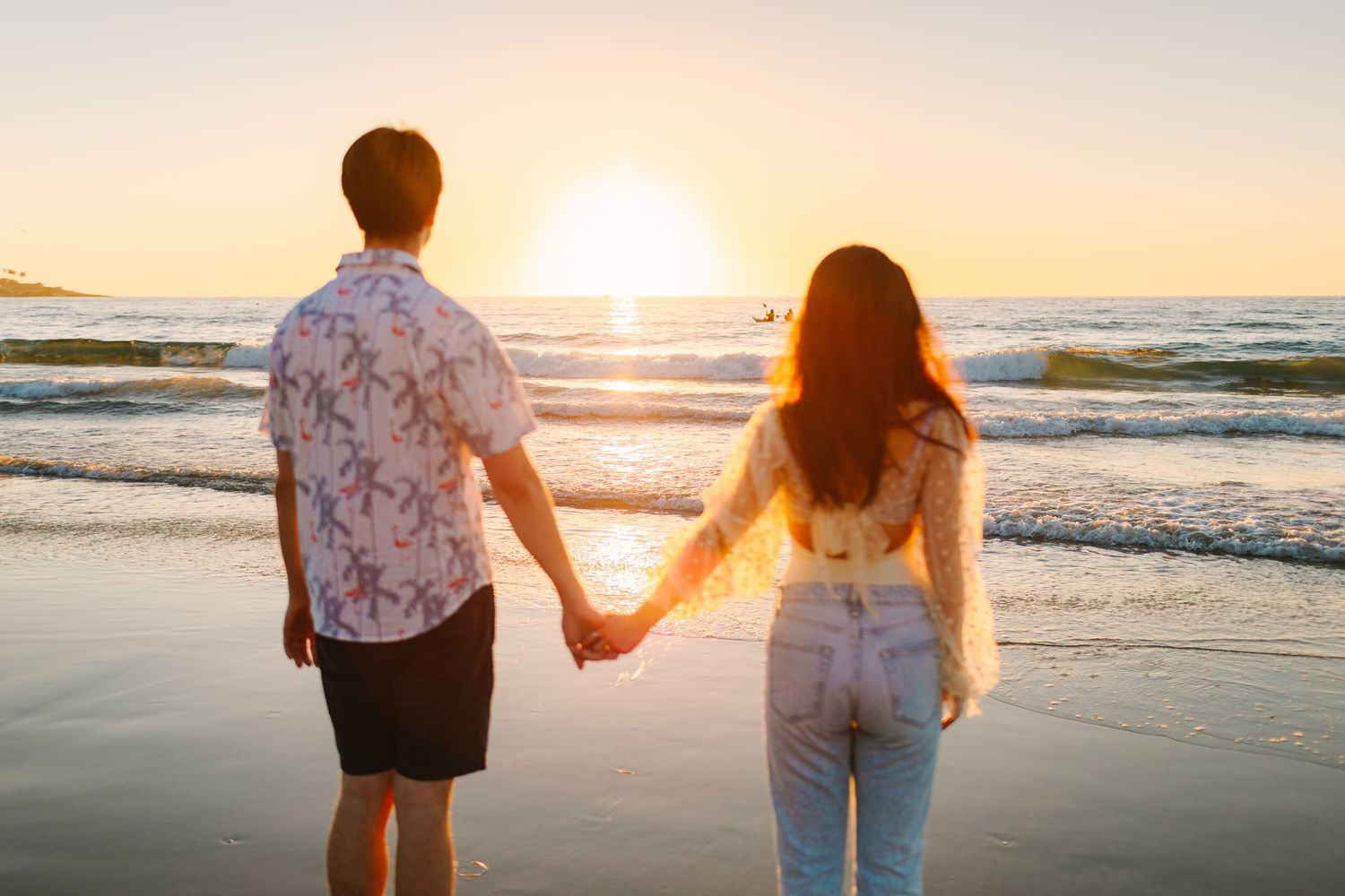 Couple holding hands while watching sunset together on a peaceful beach with waves rolling in.