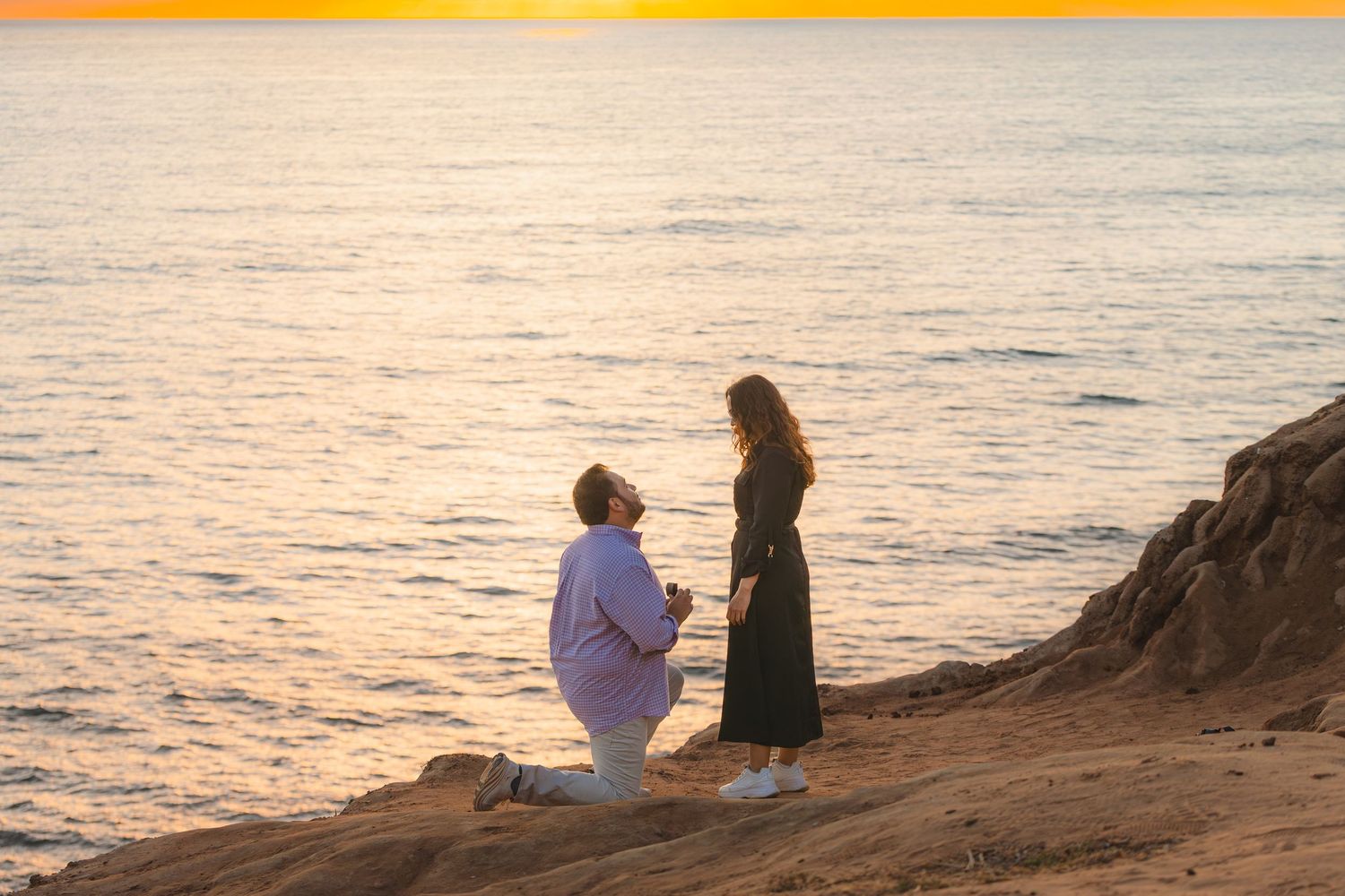 Two people sit on a rocky cliff overlooking a serene ocean at sunset, creating a peaceful silhouette against the sky.