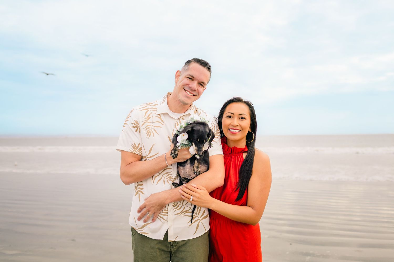A couple stands on a beach holding their small black dog while smiling on a cloudy day.