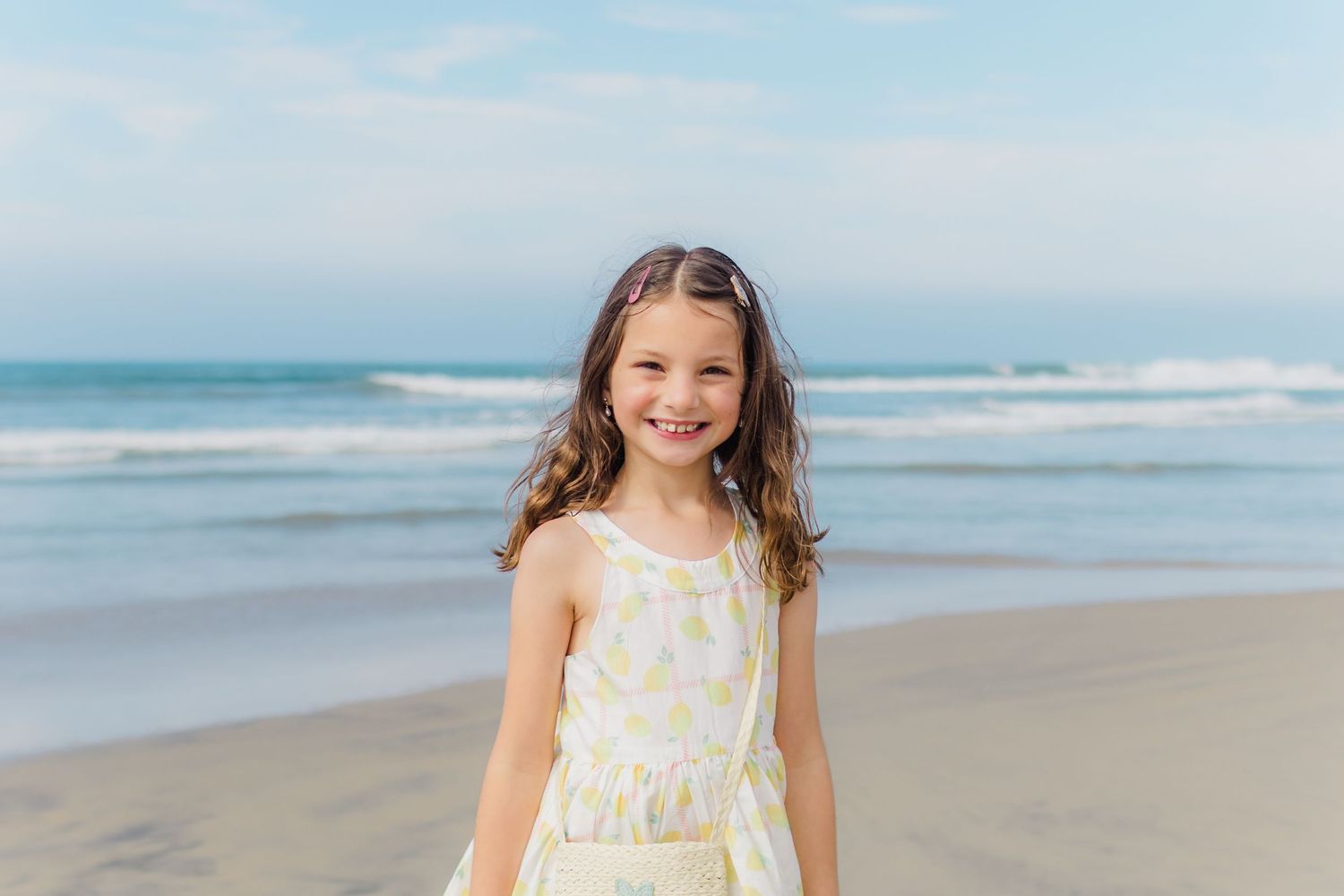 Child in white summer dress stands smiling on sunny beach with ocean waves in background.