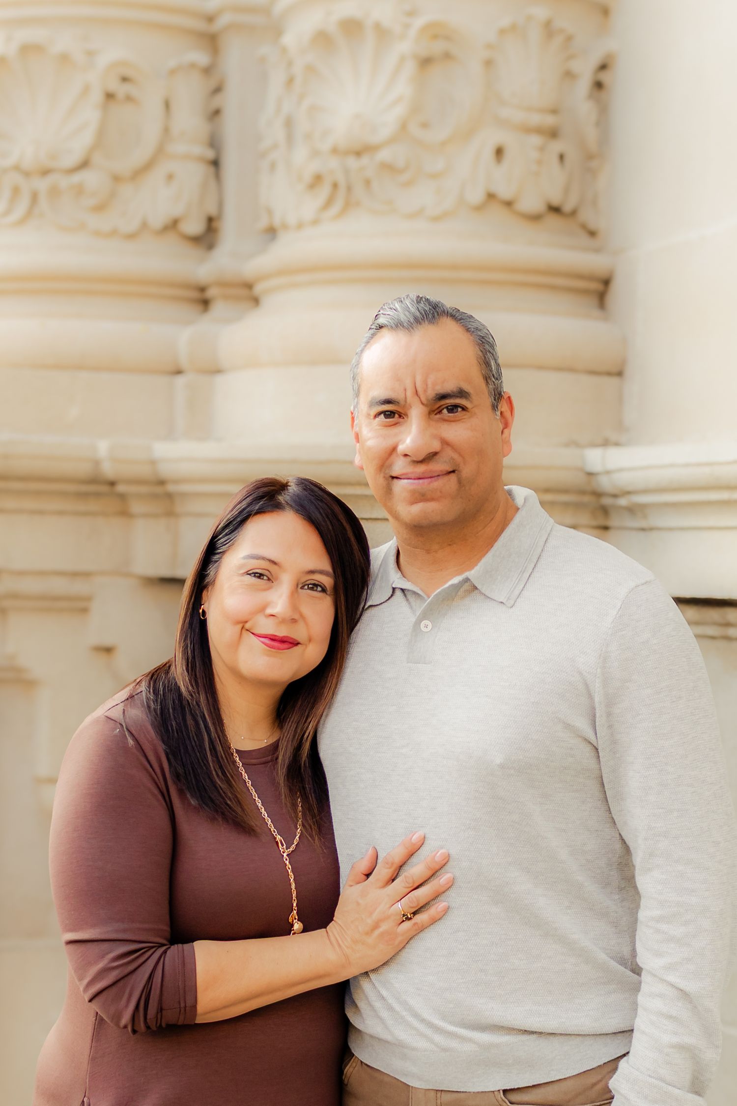 Two people pose together against ornate stone columns wearing casual attire and warm smiles.