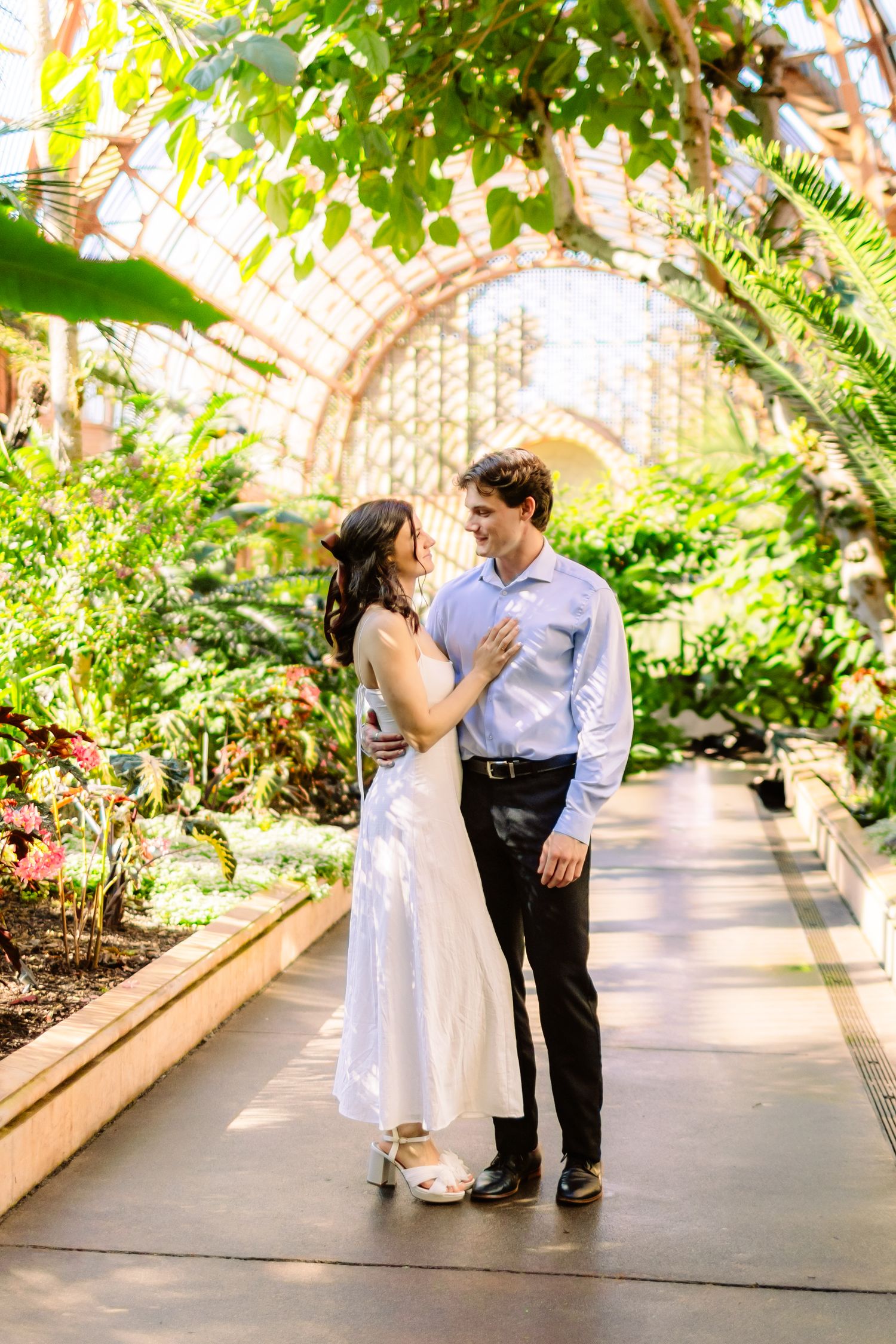 Couple dancing together in a sunlit greenhouse pathway lined with tropical foliage and latticed windows.