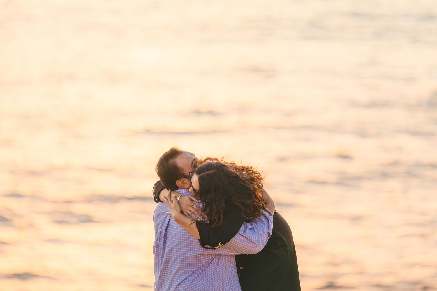 Two people share a tender embrace at sunset with golden light reflecting on the water in the background.