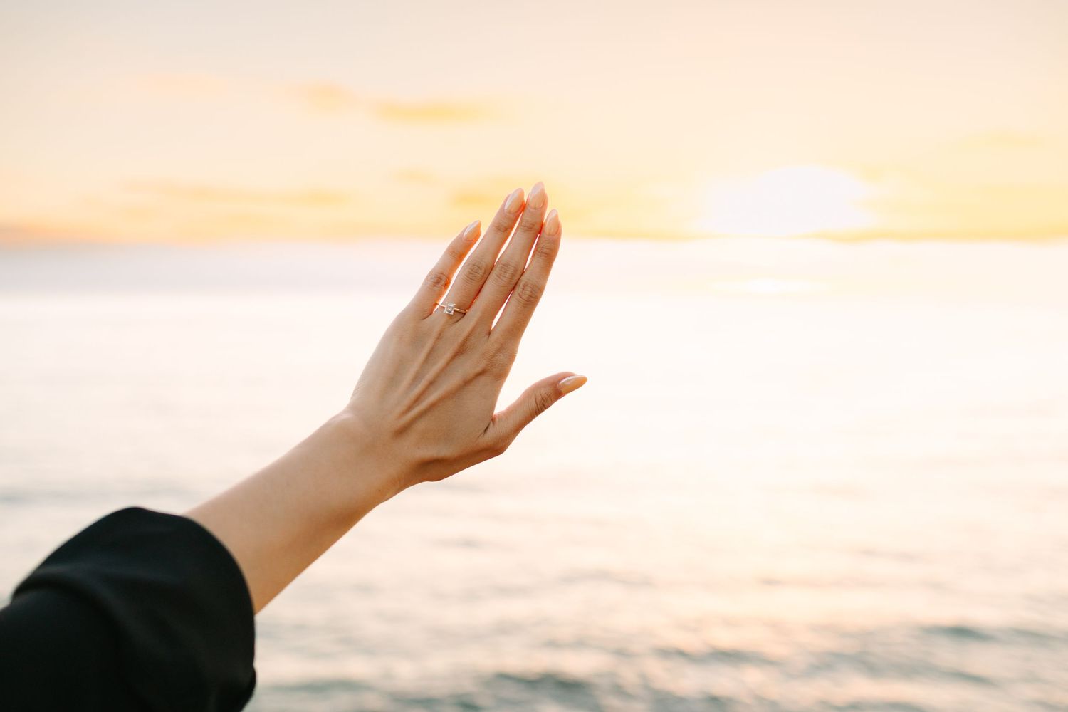 Hand reaching towards warm sunset sky over ocean horizon creating silhouette against golden light.