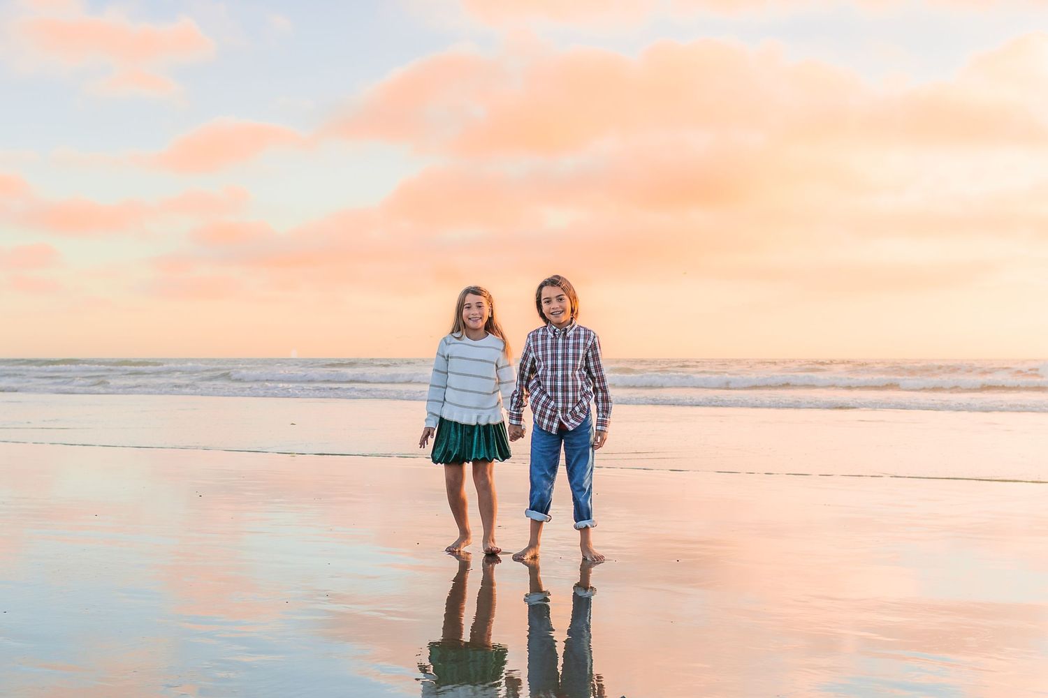 Two people stand together on a serene beach at sunset with pink and orange sky reflected in the wet sand.